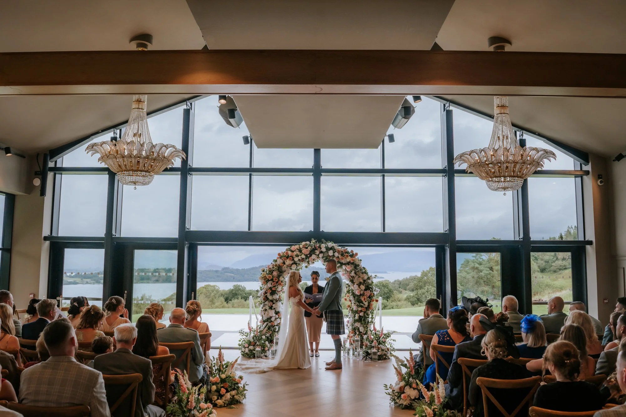 A wedding ceremony taking place indoors in front of large glass windows overlooking a lake and hills. The bride and groom stand under a floral arch with an officiant, surrounded by seated guests. The venue features chandeliers and elegant decor.