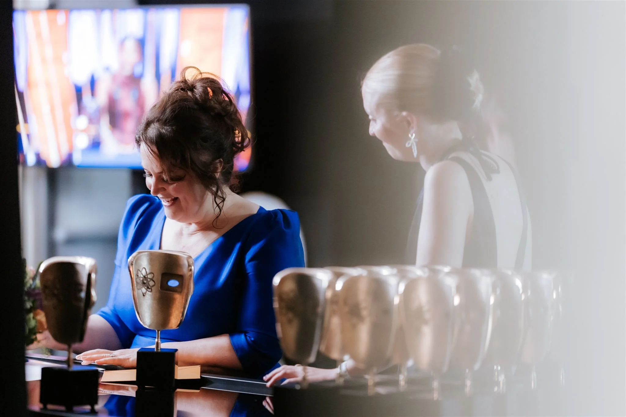 Two women in formal attire are smiling, one holding a gold-colored award; there are multiple awards on the table in front of them, with a TV screen showing blurred images in the background.