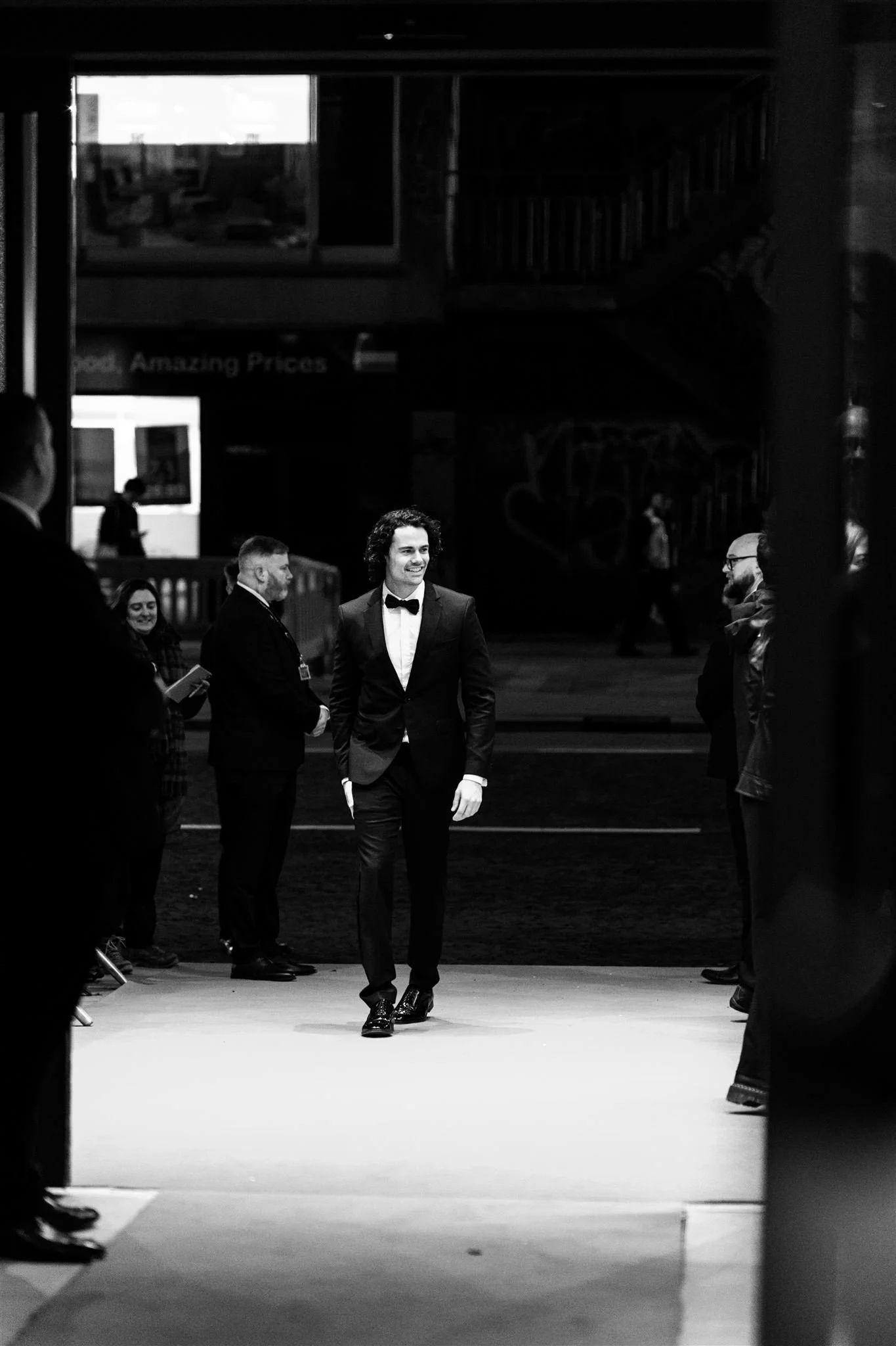 A man in a tuxedo walking on a red carpet surrounded by people in formal attire at an event, viewed through a partially open door.