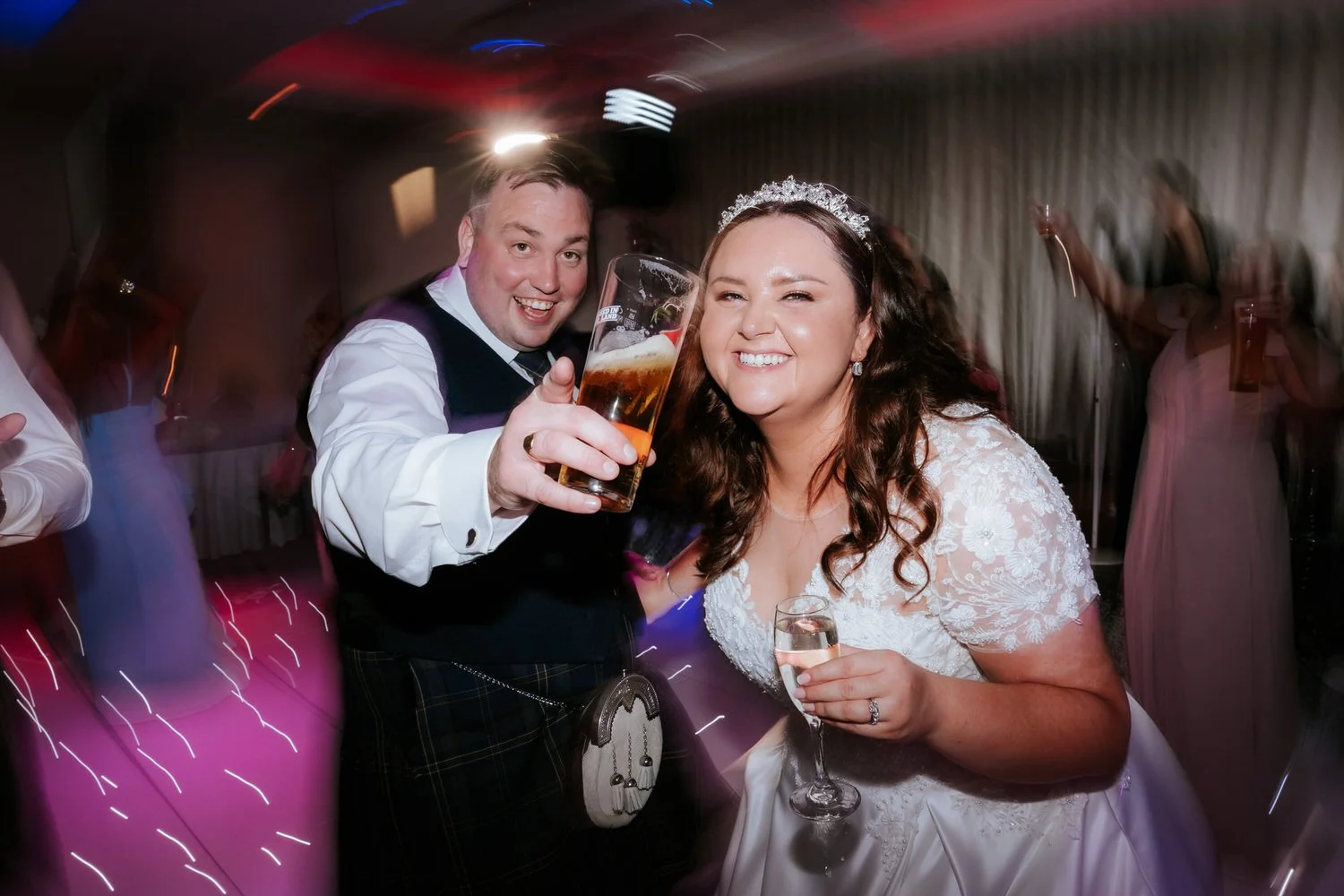 Bride and groom smiling and raising drinks at wedding reception, bride wearing white lace dress and tiara, groom in vest and kilt, party guests in the background.