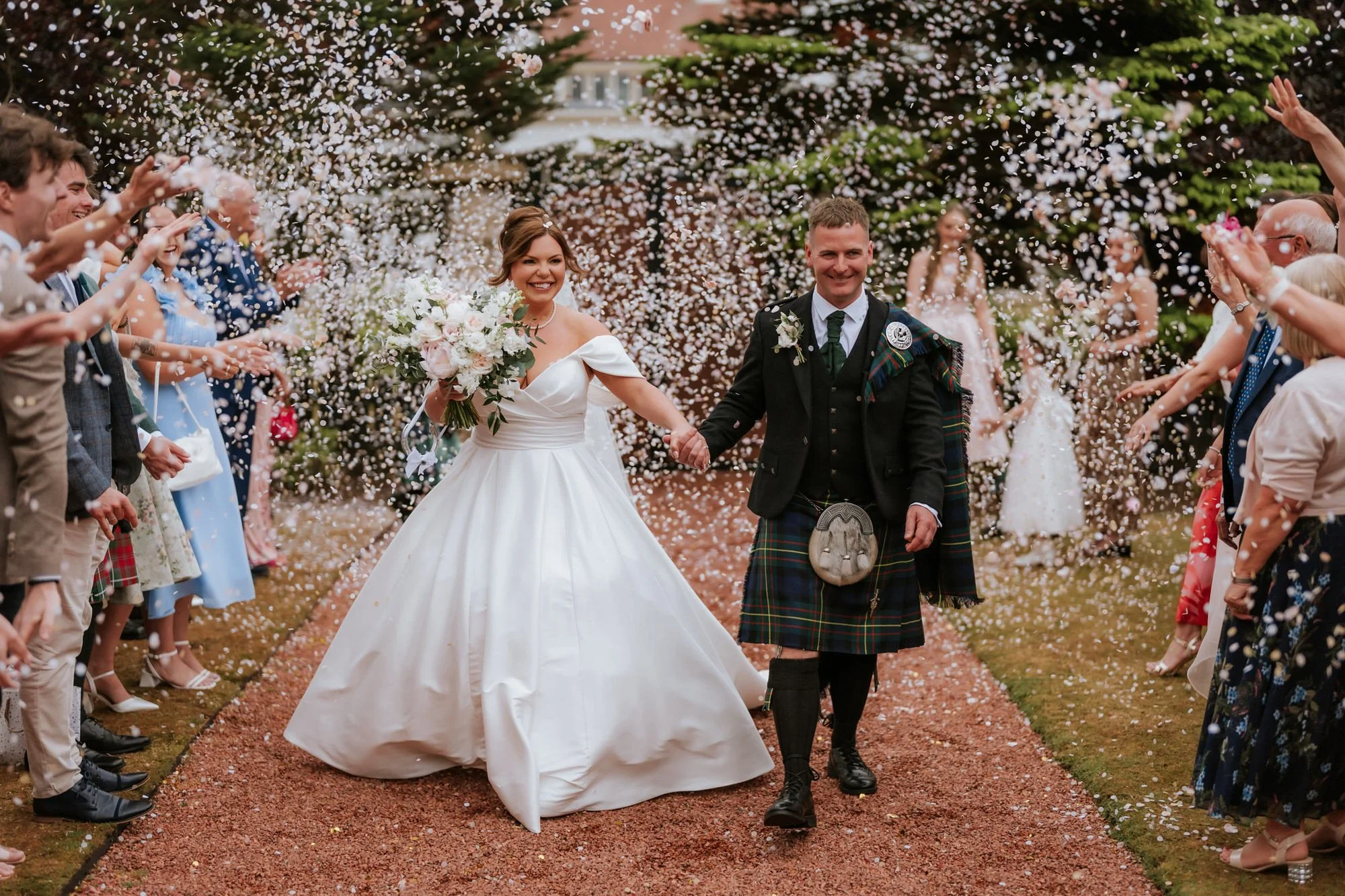 Bride and groom walking hand in hand through guest shower of rice or confetti at wedding ceremony outside, surrounded by friends and family.