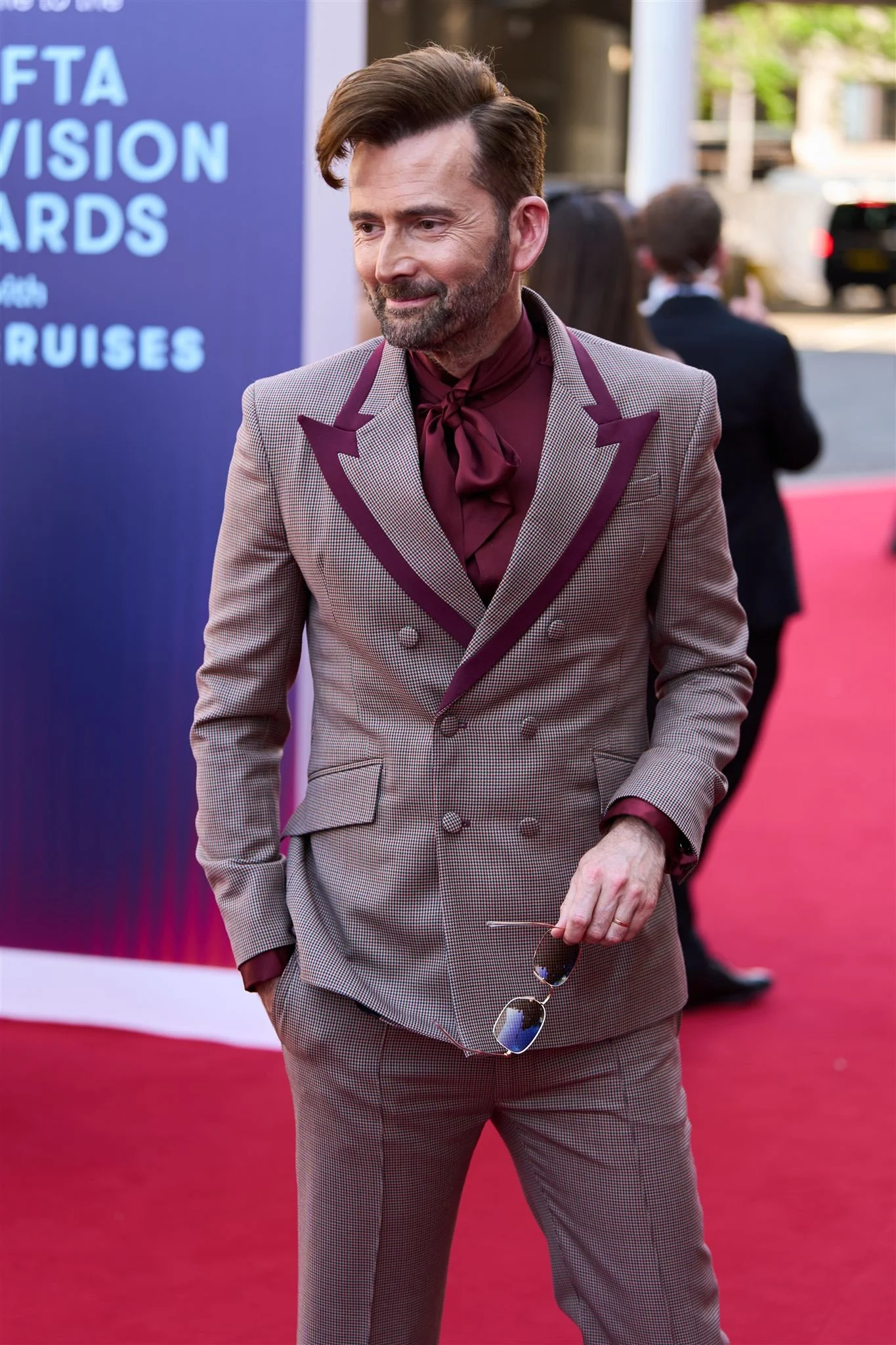 A man on a red carpet wearing a double-breasted checkered suit with purple accents, holding sunglasses.