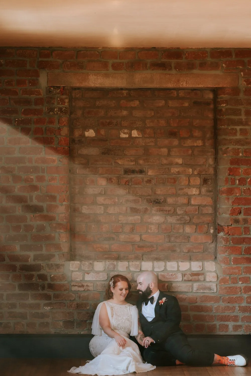 Couple in wedding attire sitting on the floor against a brick wall.