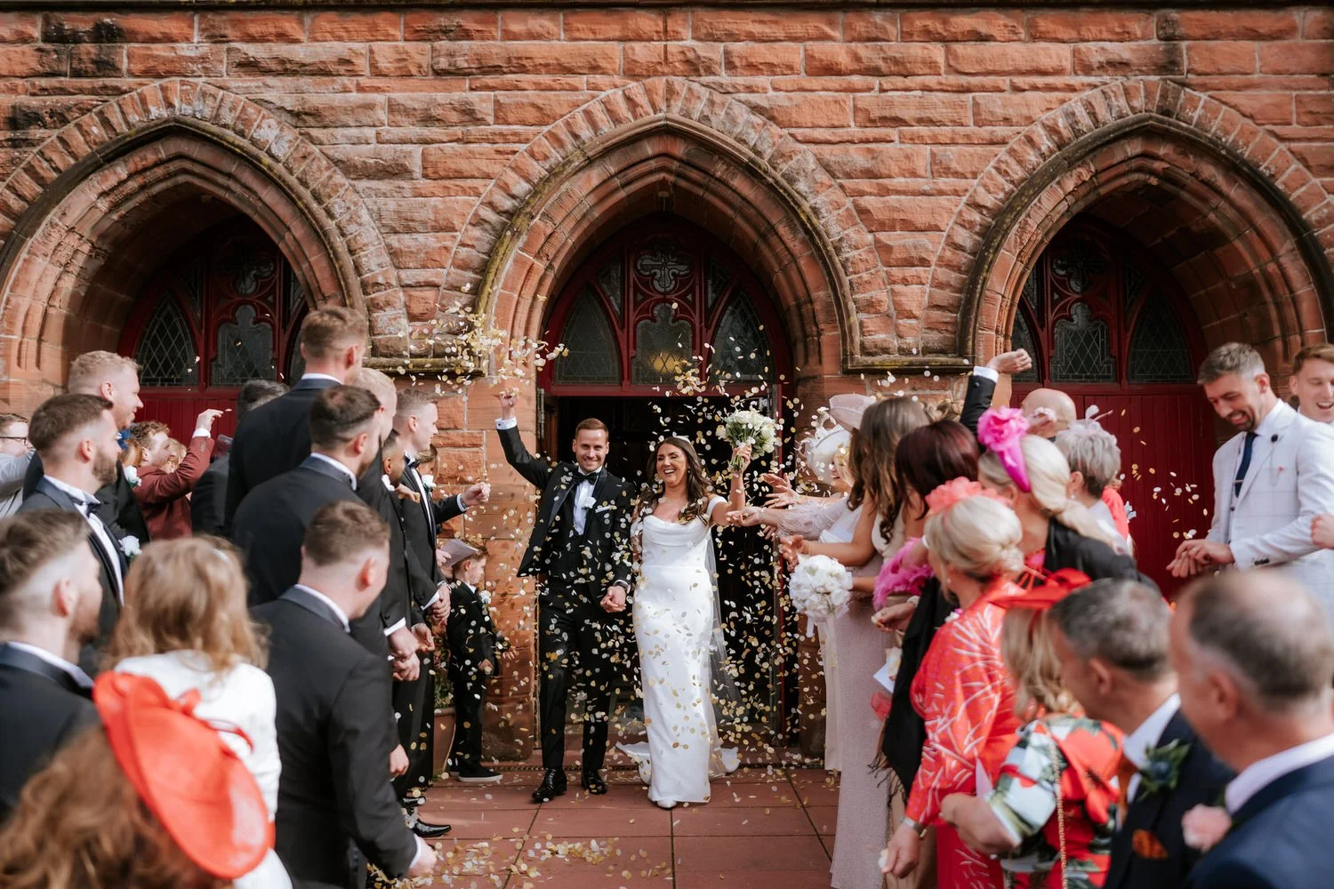 A newlywed couple walking out of a church, surrounded by wedding guests throwing confetti, with a red brick church in the background.