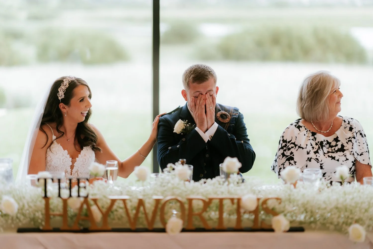 A bride with a white wedding dress and a headband is seated at a table, smiling and touching the groom's shoulder. The groom in a military uniform is covering his face with his hands, appearing emotional. An older woman in a black and white floral dr
