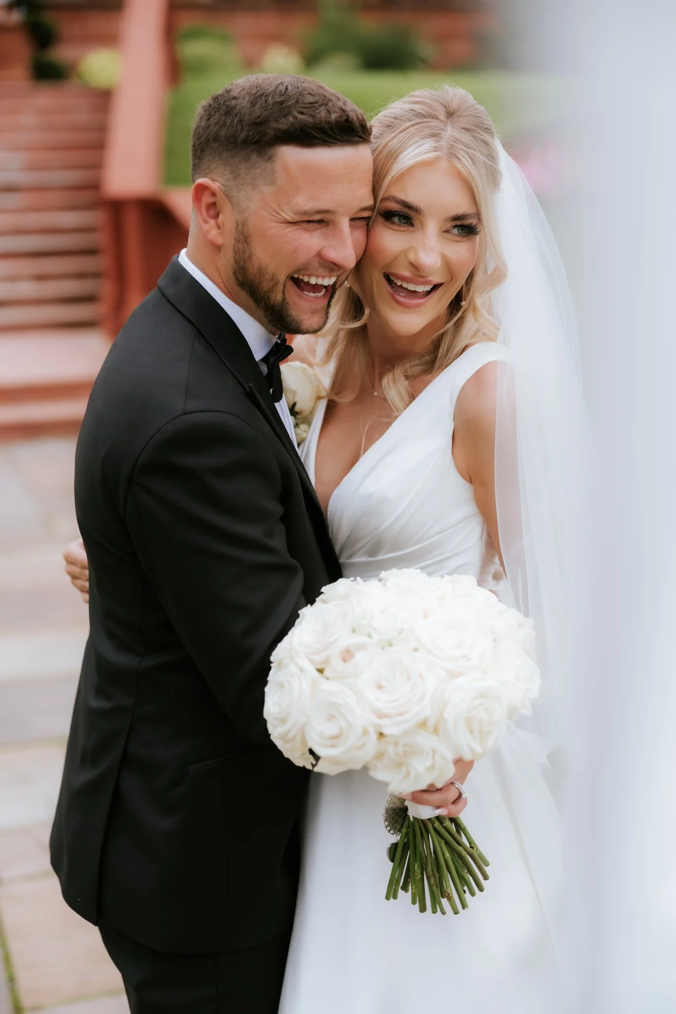A bride and groom on their wedding day, smiling and embracing each other, with the bride holding a bouquet of white roses.