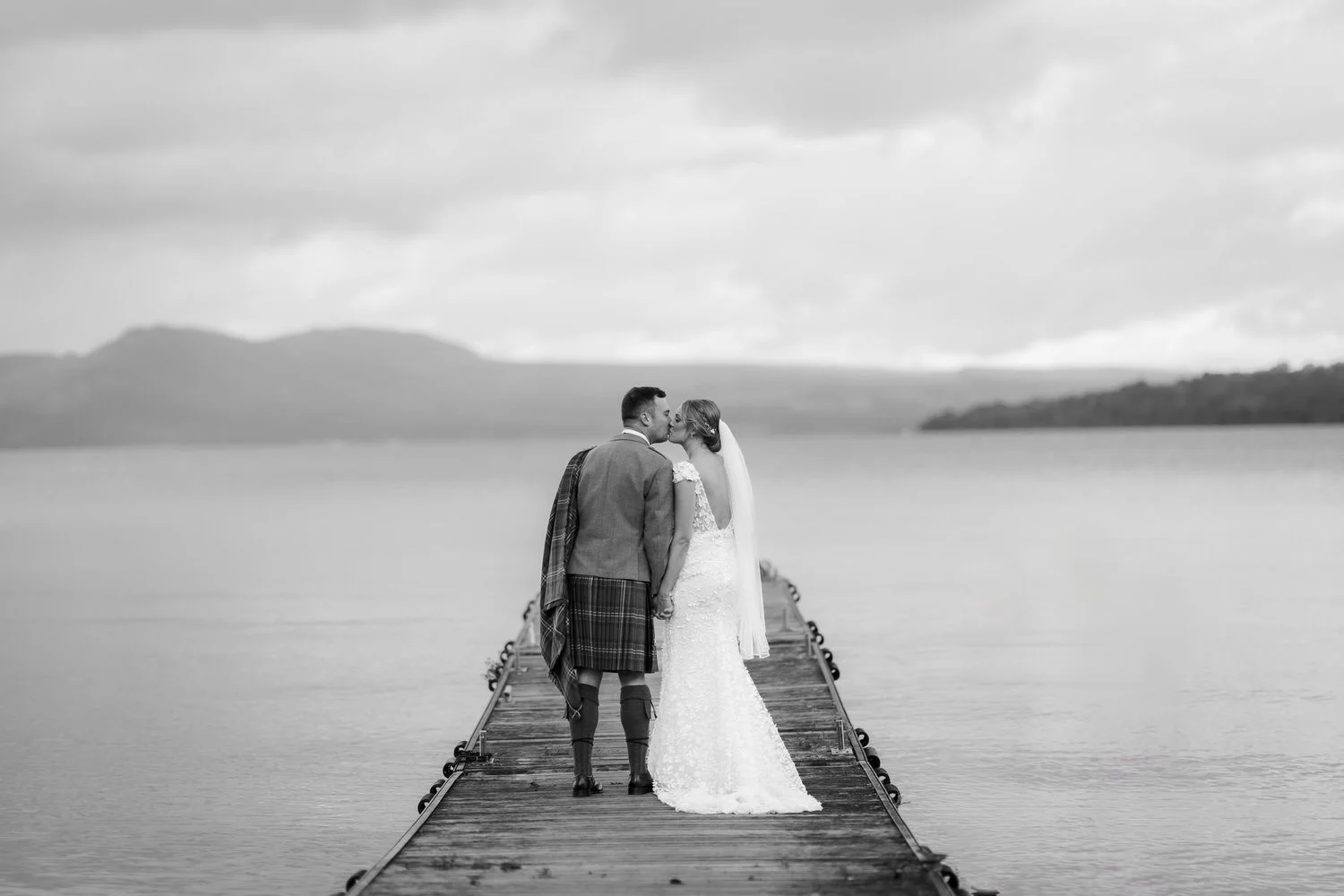 Black and white photo of a newlywed couple kissing on a wooden dock by a lake, with mountains in the background.