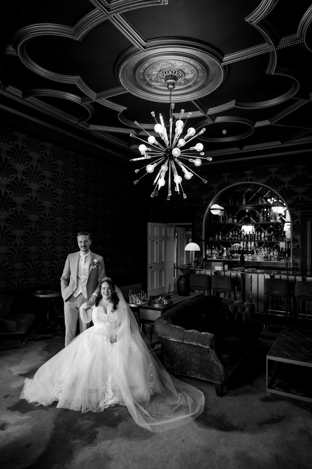 Black and white photo of a newlywed couple in an elegant, vintage-style room, with a chandelier hanging from decorated ceiling, bar area in the background, and ornate wallpaper. The bride is sitting in a wedding gown with a veil and tiara, smiling, w