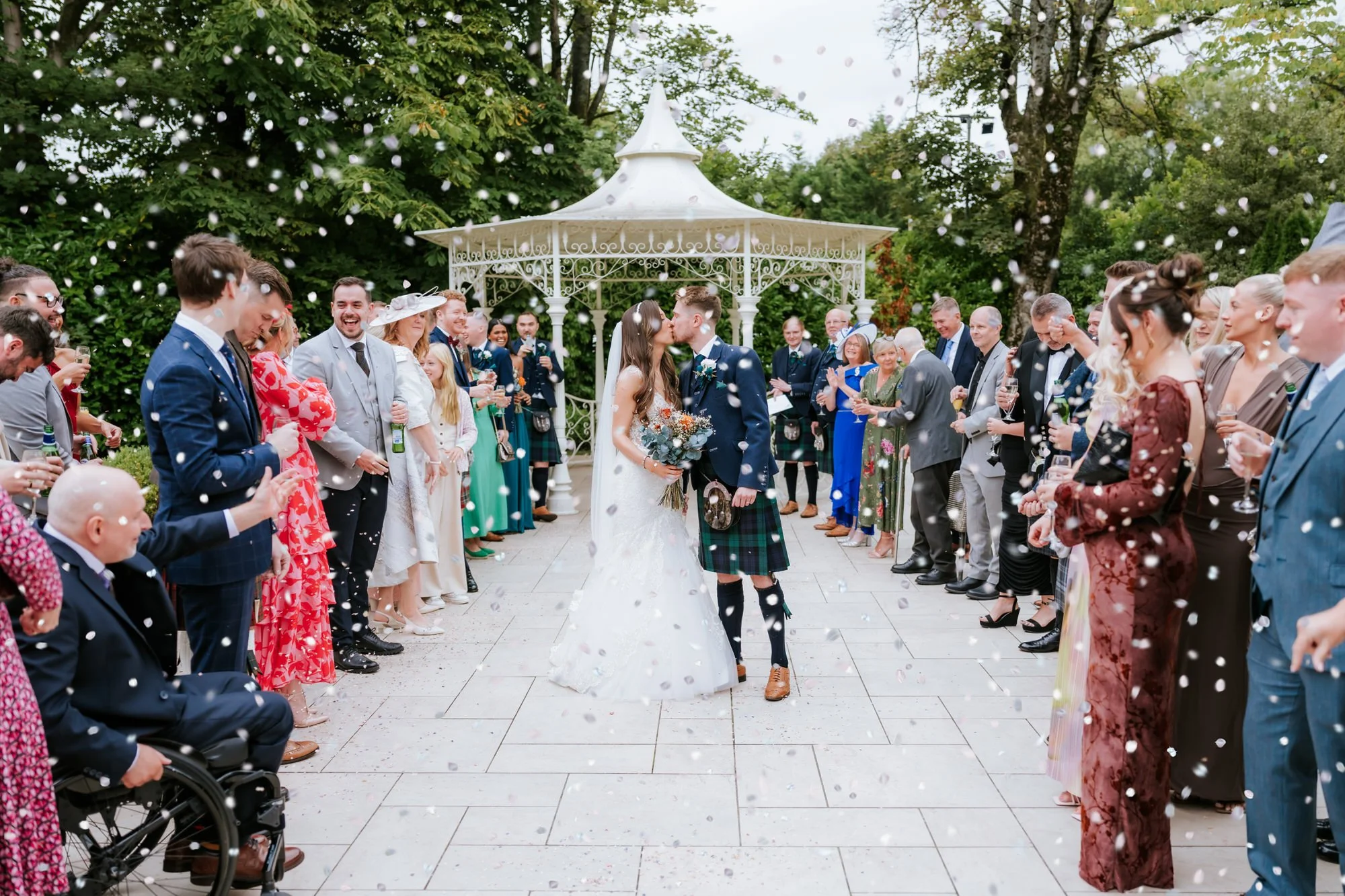 A bride and groom kissing under a gazebo at their wedding, surrounded by guests throwing confetti outdoors.