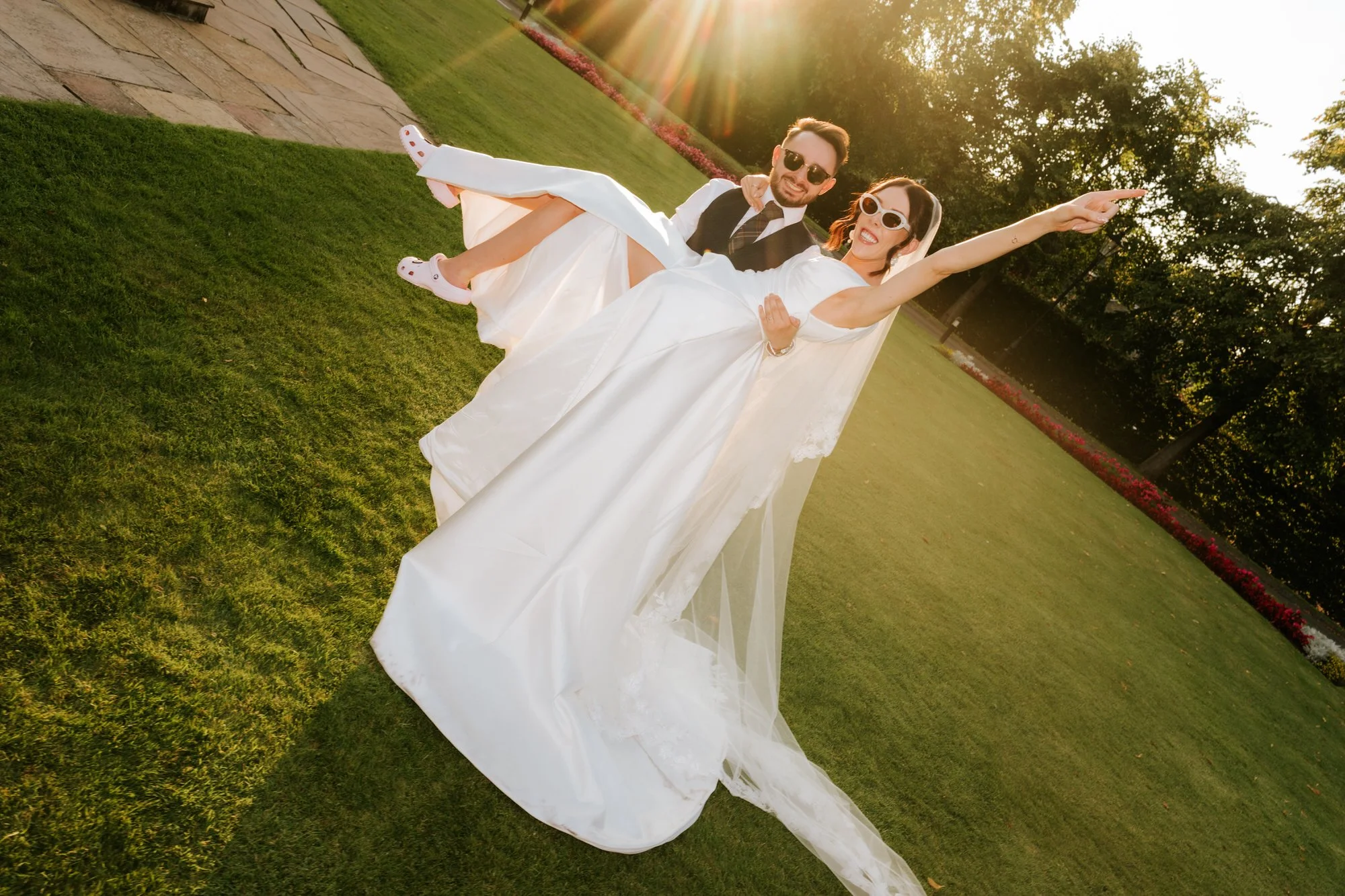 A bride and groom in wedding attire dancing outdoors on a grassy area during sunset, with trees and flowers in the background.