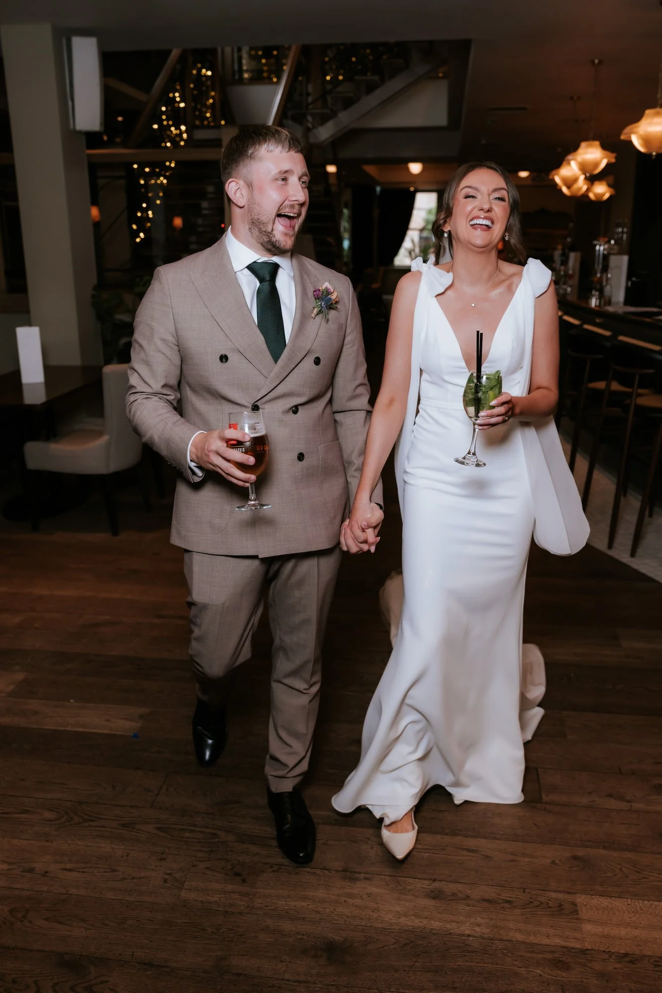 A smiling couple, the man in a tan suit and the woman in a white gown, holding hands and clinking drinks at a wedding reception.