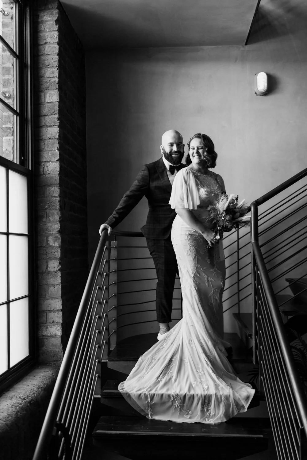 Black and white photo of a newlywed couple standing on a staircase indoors. The woman is holding a bouquet and wearing a lace wedding gown, while the man is in a suit and bowtie, smiling.