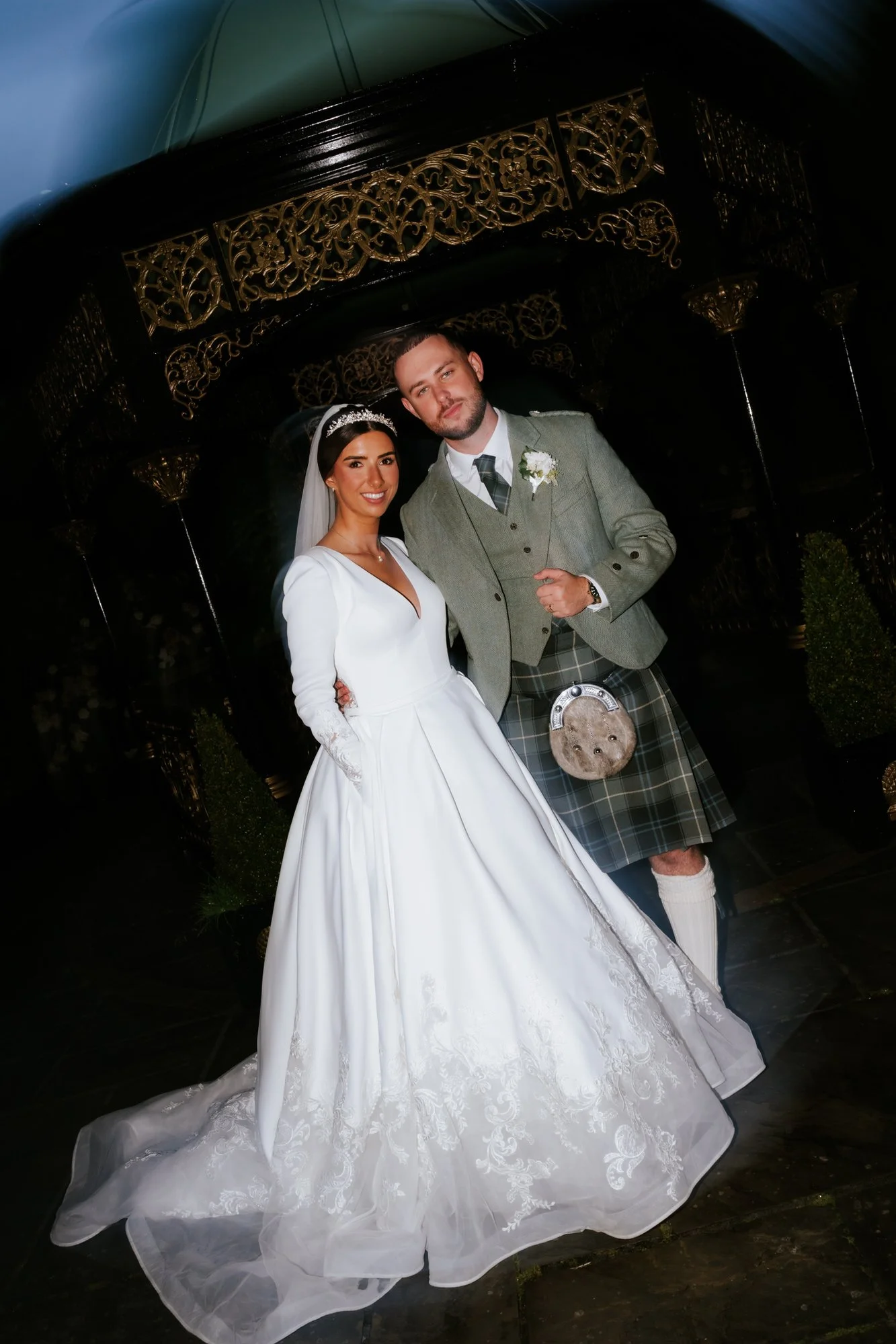 Bride and groom dressed in traditional wedding attire, standing together outdoors at night, with decorative black and gold structure behind them.