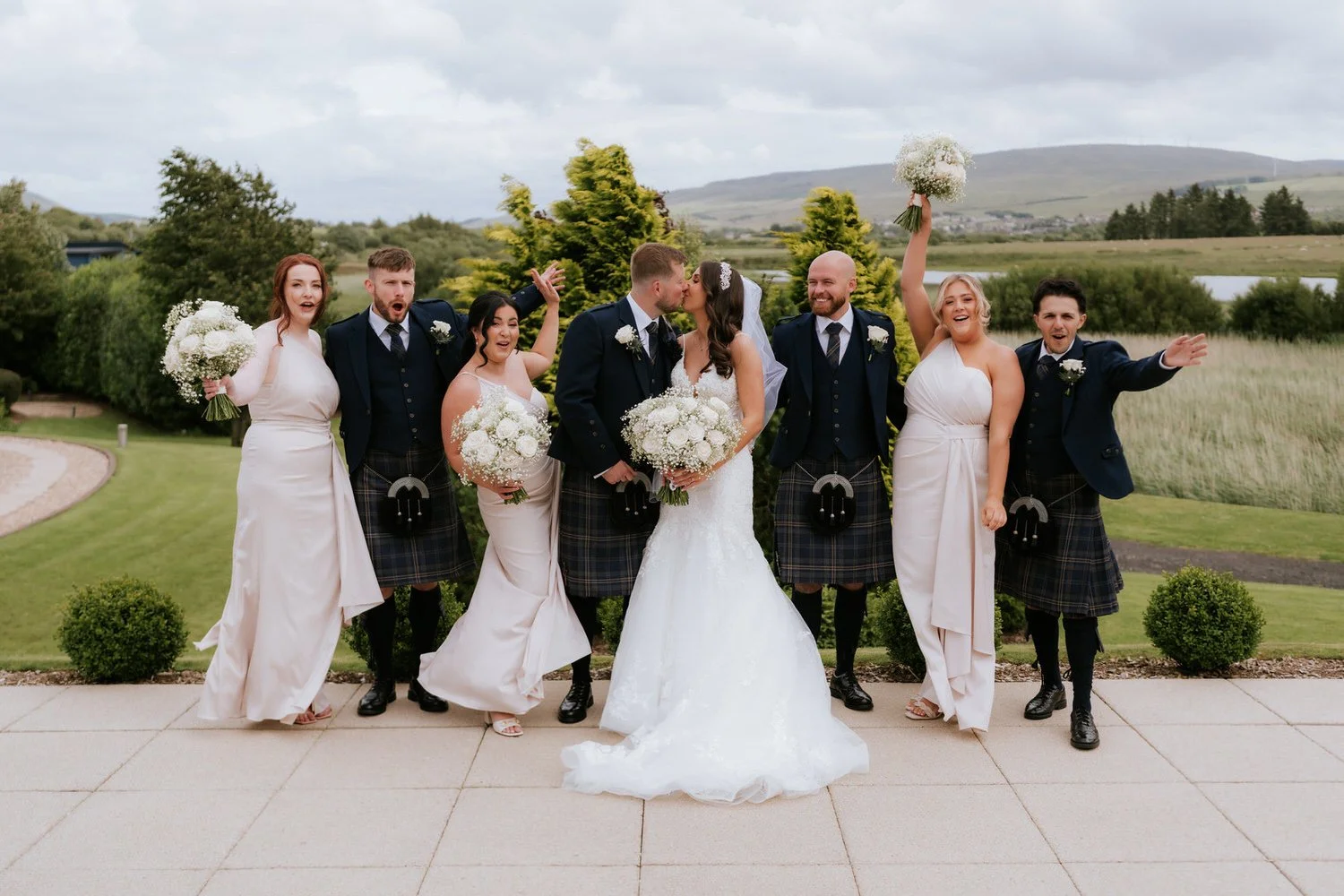 Wedding party celebrating outdoors, with bride and groom kissing in the center, surrounded by bridesmaids and groomsmen, some in kilts, holding bouquets, on a lush green lawn with trees and hills in the background.