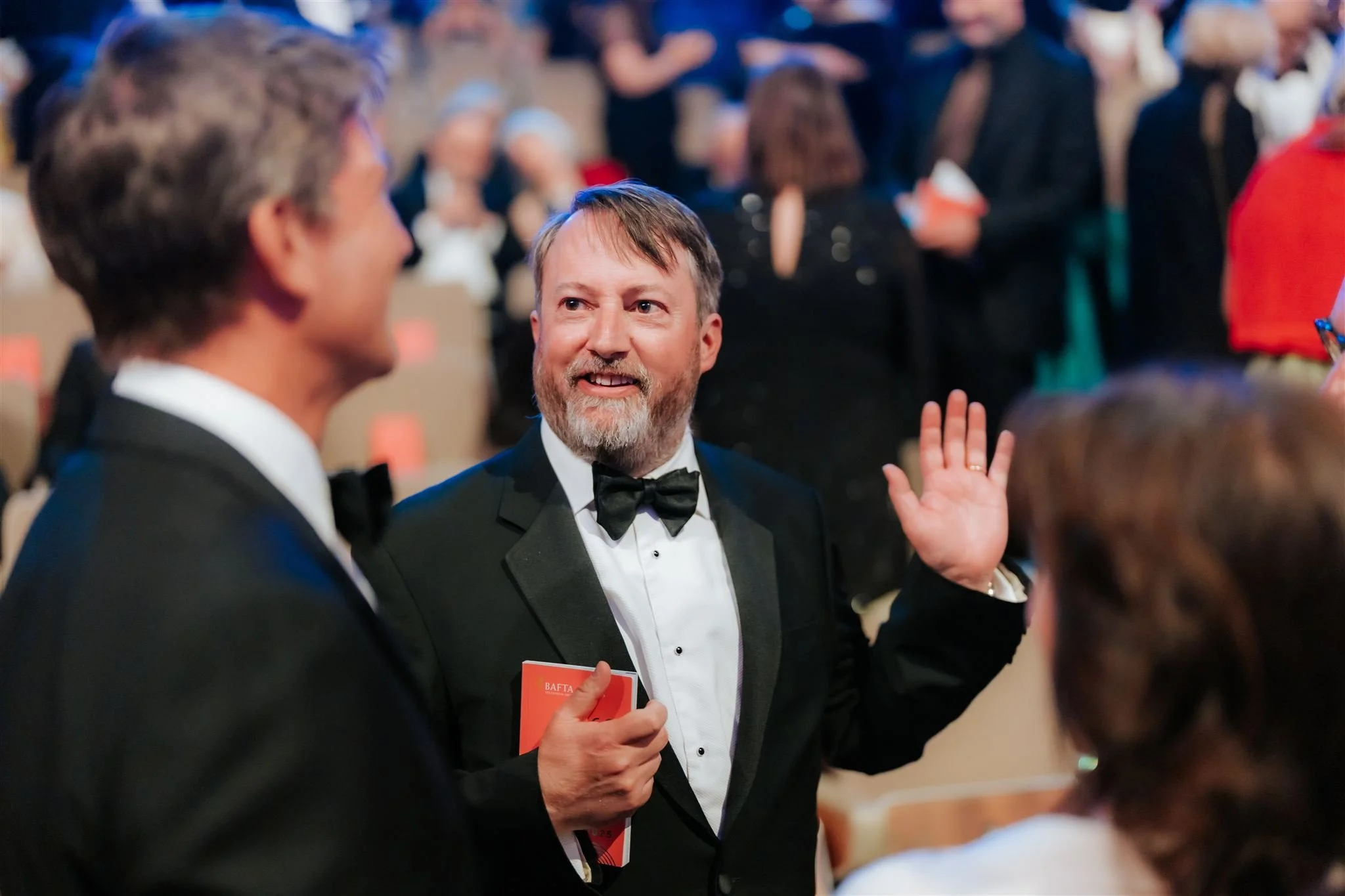 Man with gray beard wearing a tuxedo and bow tie, waving and smiling, at a formal event with others in tuxedos and evening gowns in the background.