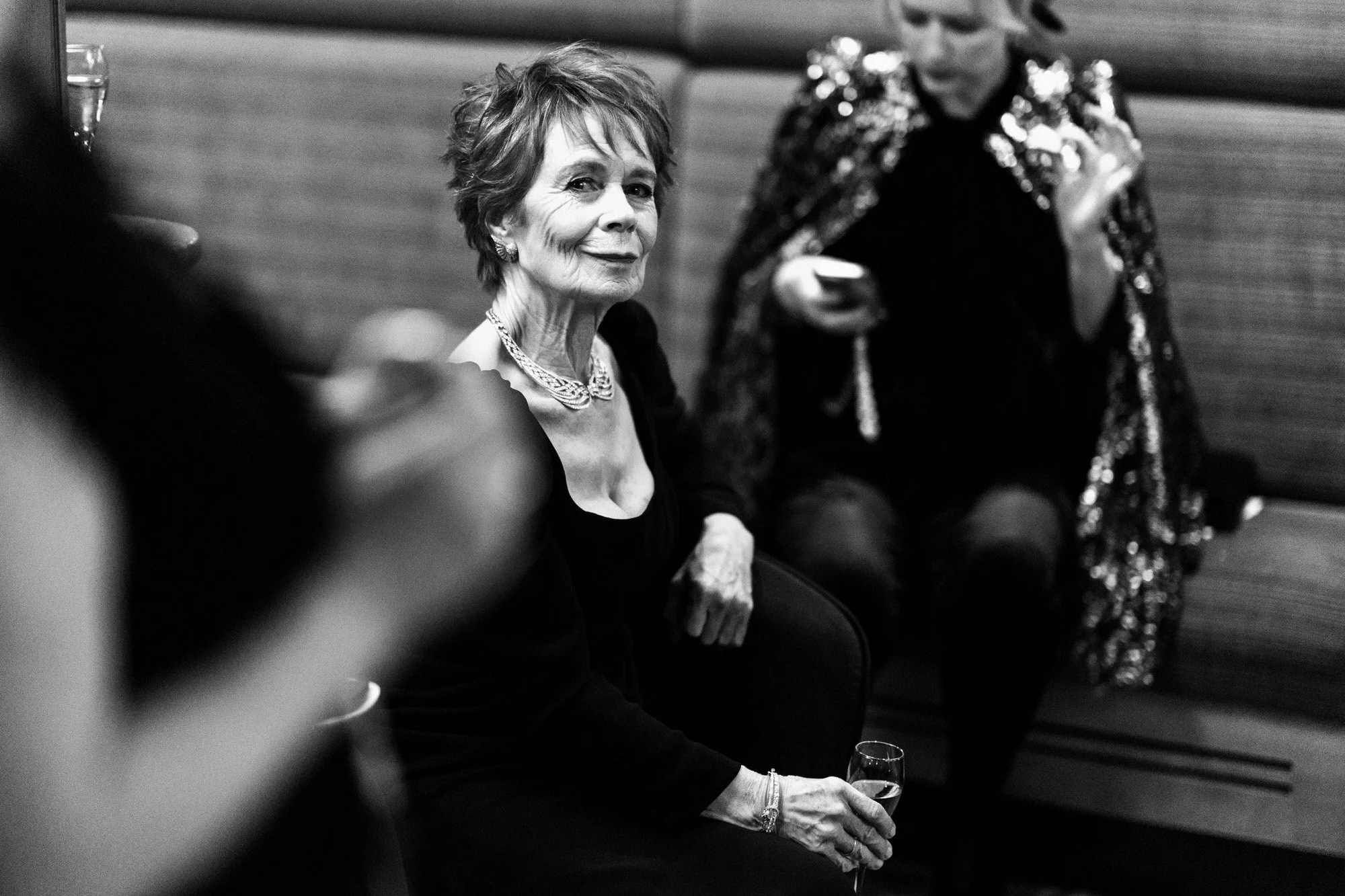 Celia Imrie seated with a drink in the green room at the 2025 BAFTA Film Awards, photographed in black and white.