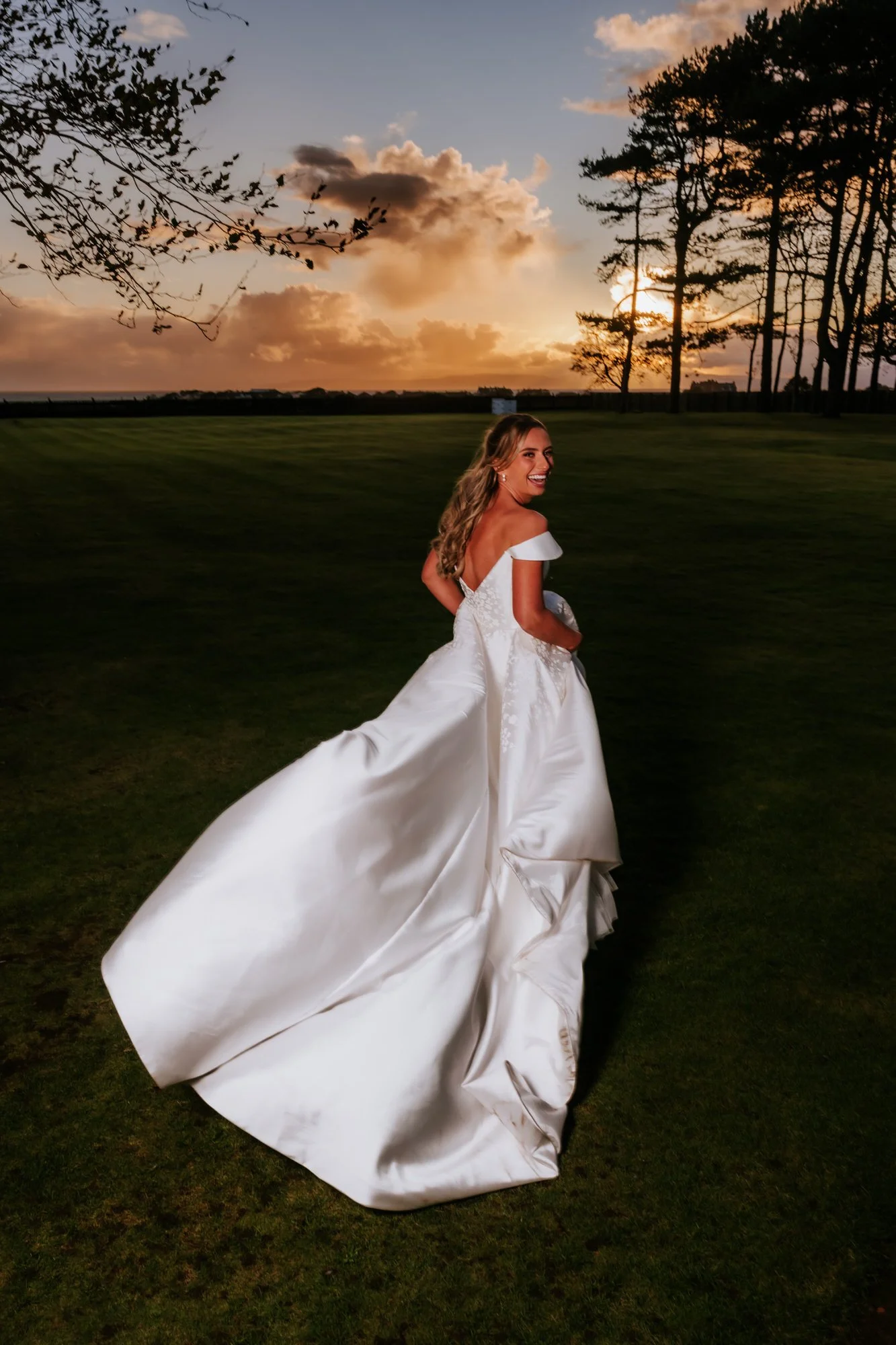 A woman in a white wedding gown walking outside in a grassy area during sunset with trees and clouds in the background.