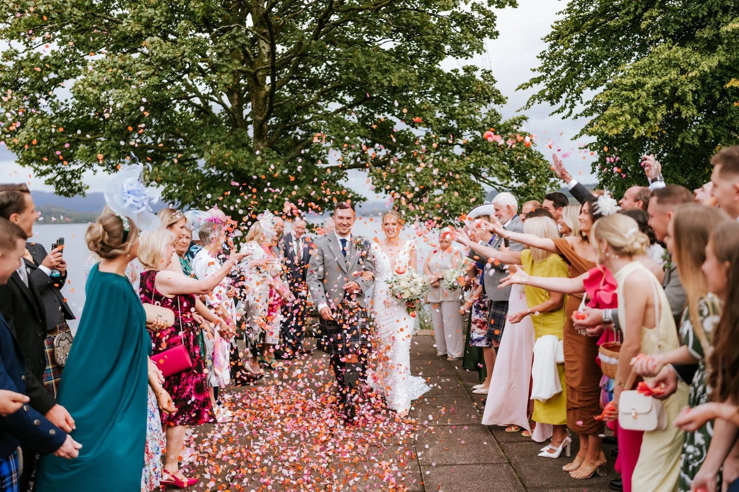 Bride and groom walking together while wedding guests celebrate and throw colorful confetti outdoors.