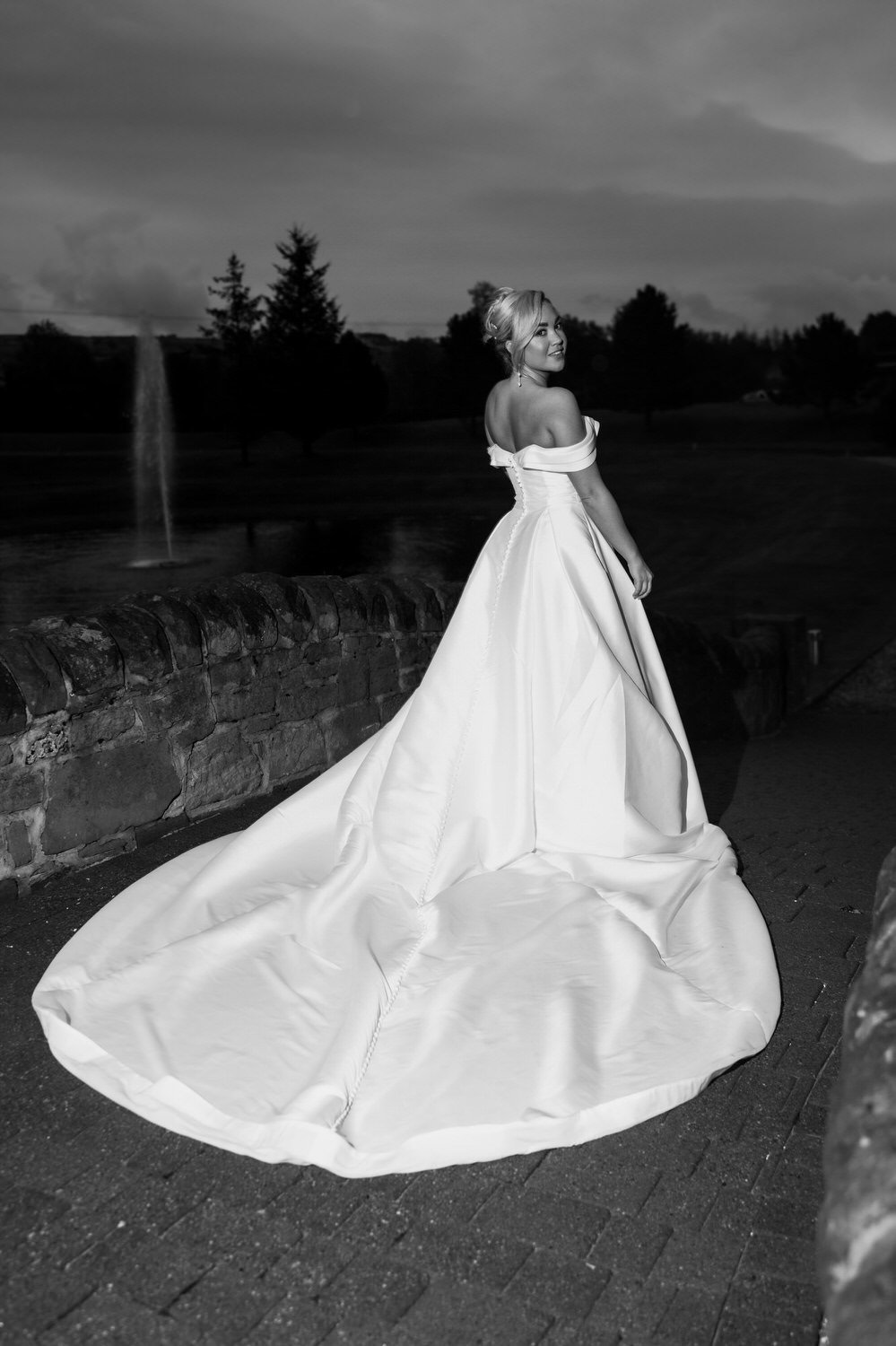 A woman in a strapless wedding gown with a long train standing on a stone pathway near a brick wall, outdoors in the evening with trees and a fountain in the background under a cloudy sky.