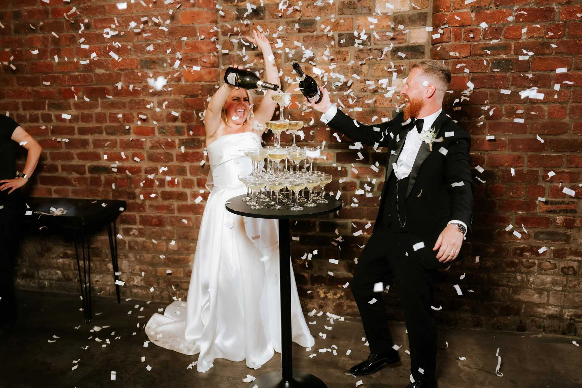 Bride and groom celebrating with champagne over a tower of glasses, surrounded by falling confetti against a brick wall background.