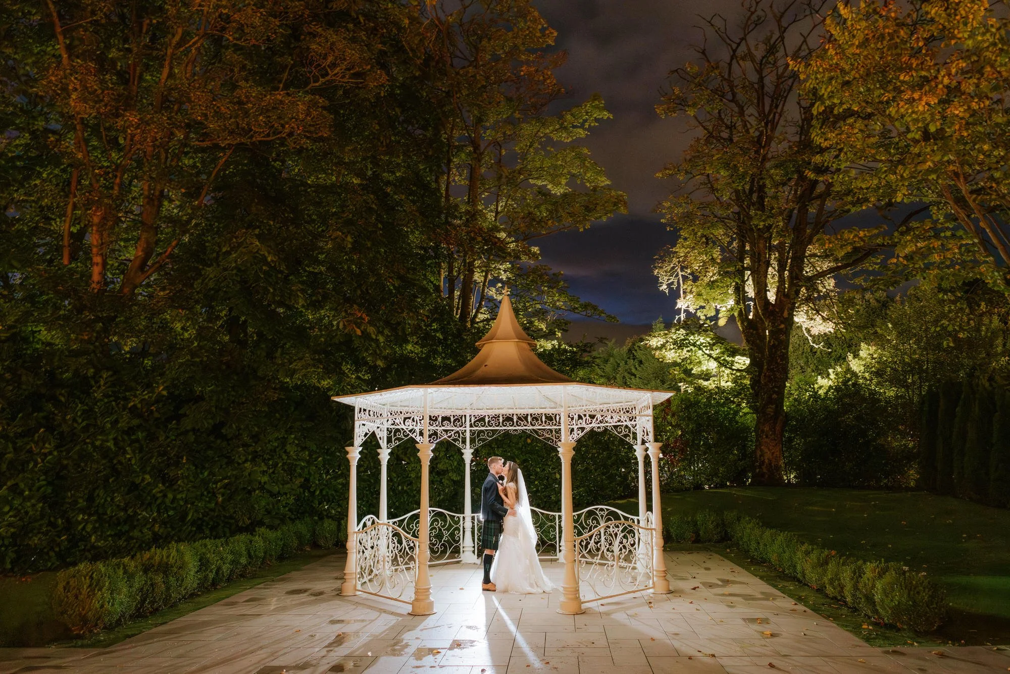 A bride and groom kissing inside a white gazebo at night, surrounded by trees and illuminated by outdoor lighting.