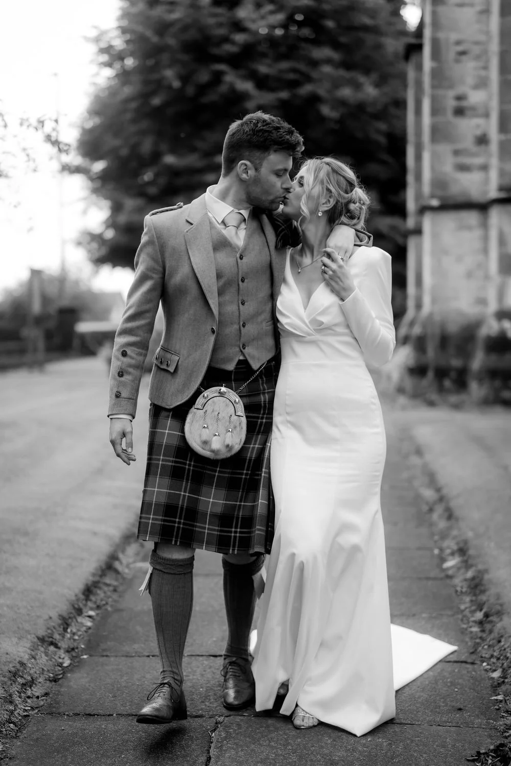 A black and white photo of a couple kissing, the man wearing a suit jacket, vest, kilt, and sporran, and the woman wearing a long white dress, walking together on a sidewalk.