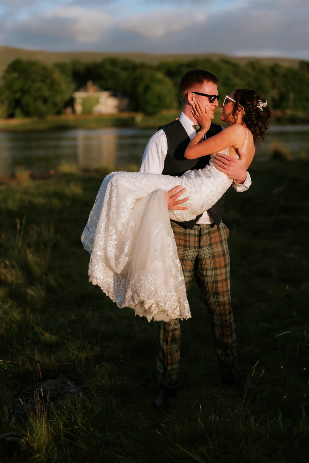 A man in plaid pants and sunglasses holding a woman in a white wedding dress and sunglasses, close together outdoors near a lake or river with green trees and hills in the background at sunset.