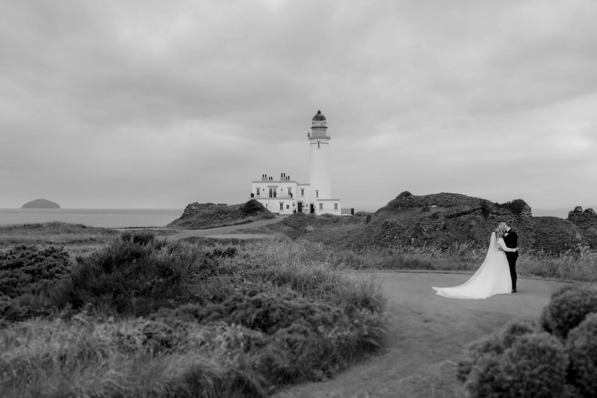 A black and white photo of a lighthouse near the coast, with a couple in wedding attire embracing in the foreground on a grassy path surrounded by bushes and rocky terrain.