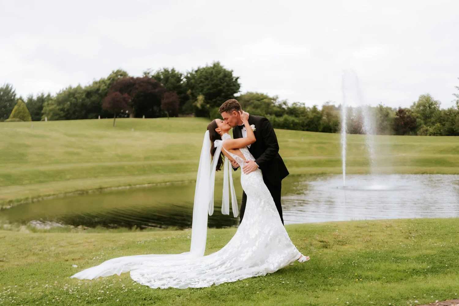 Bride and groom sharing a kiss beside a pond and fountain on a cloudy day.