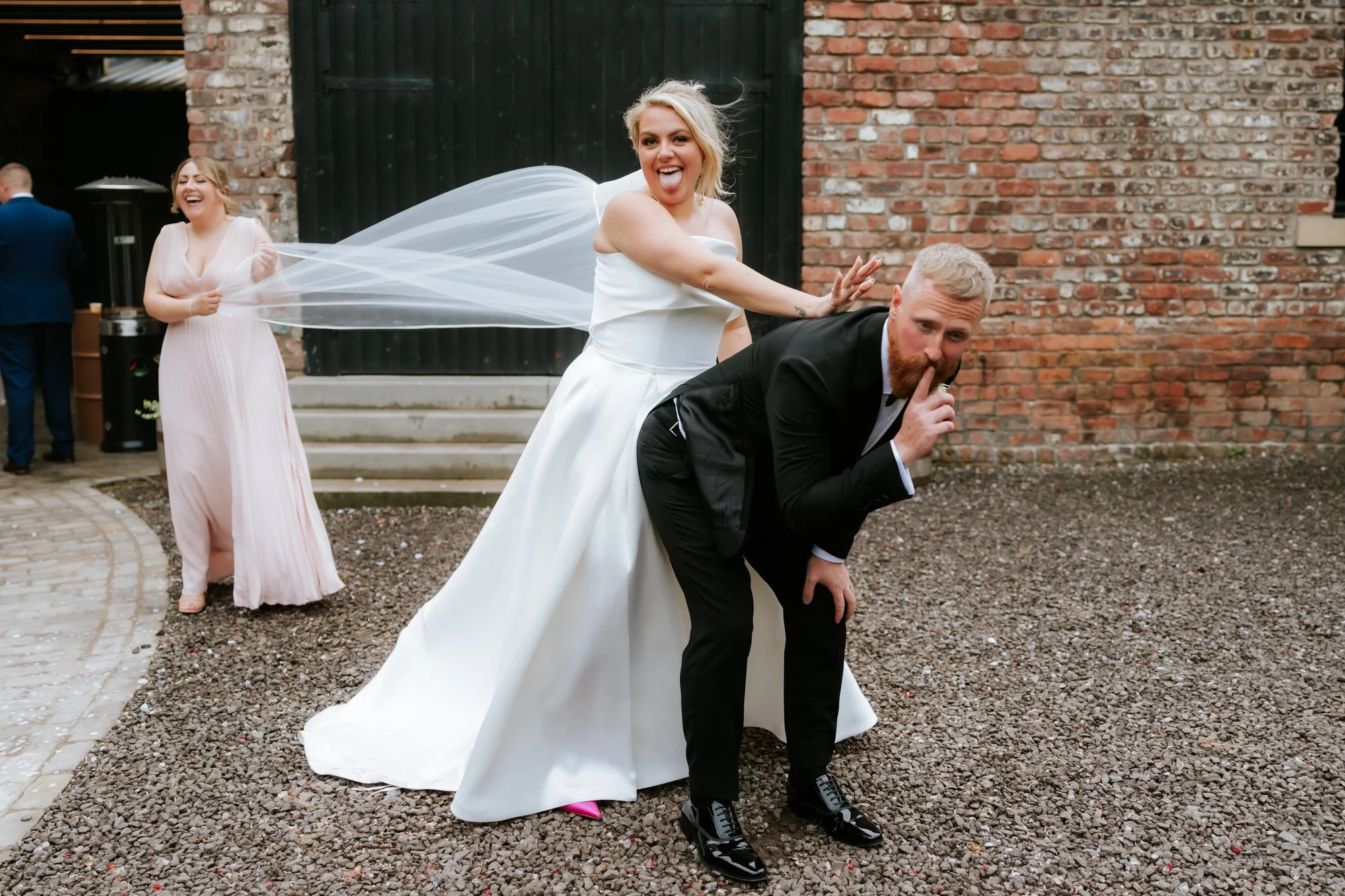 Bride playfully posing on groom's back, making a 'shh' gesture, at outdoor wedding venue with brick wall background, while bridesmaid laughs in the background.