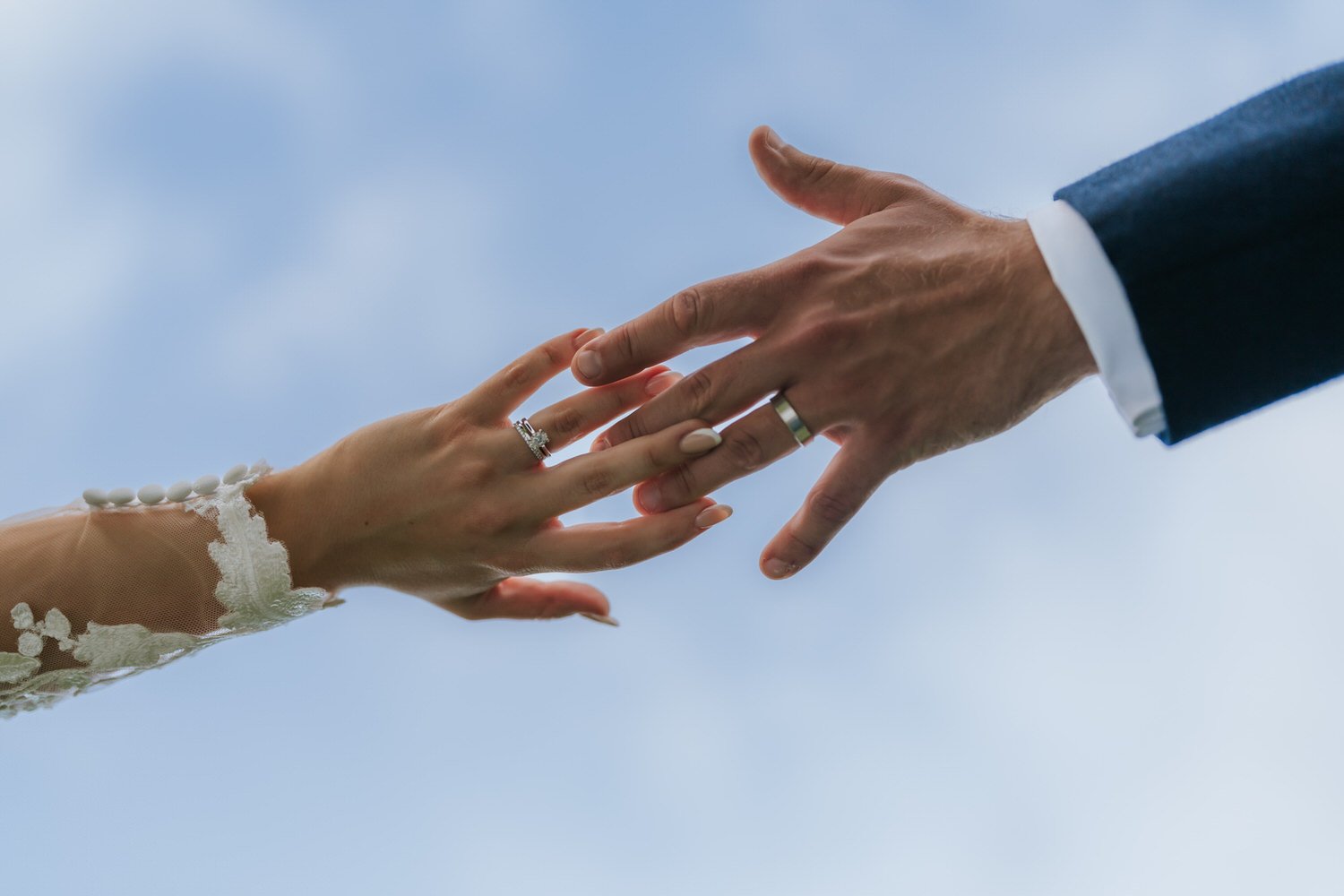 Close-up of a bride and groom reaching out to touch hands, with the bride's hand resting on the groom's fingers against a cloudy sky.