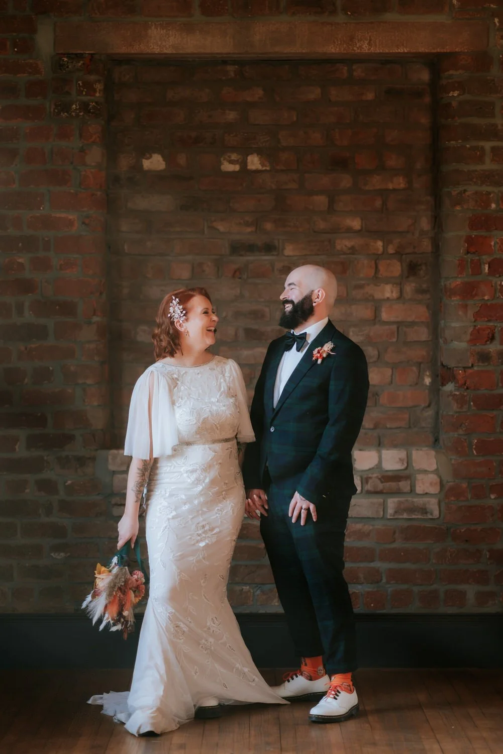 A bride and groom holding hands and smiling at each other in front of a brick wall; the bride has shoulder-length red hair and a white lace wedding dress, carrying a bouquet, and the groom has a beard and is wearing a dark plaid suit with a bow tie a