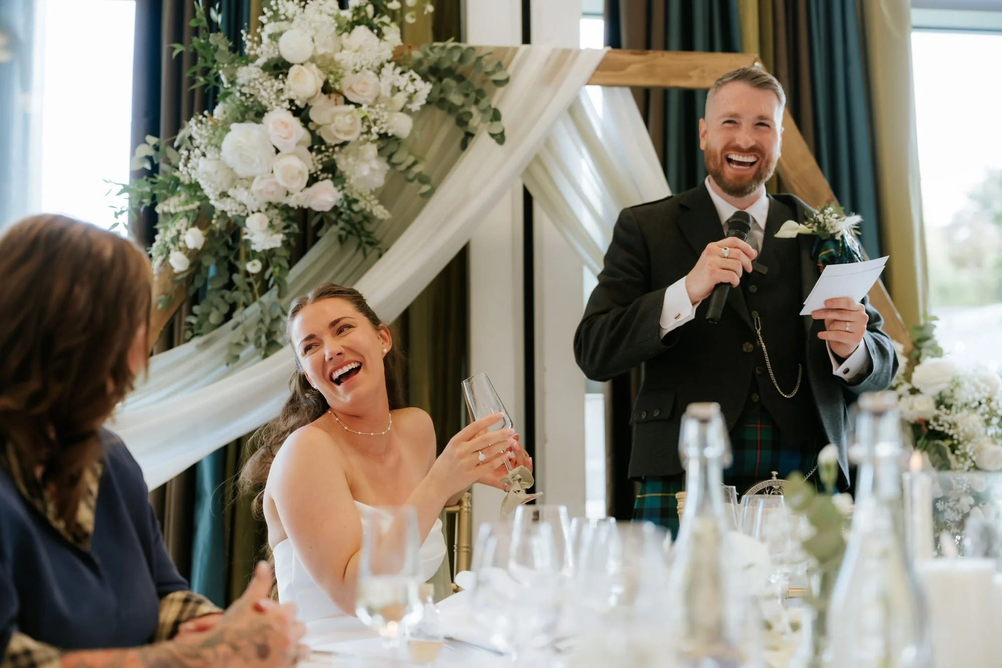 A man in a suit giving a speech at a wedding reception, with a woman smiling and laughing, sitting at a decorated table with glasses and bottles.