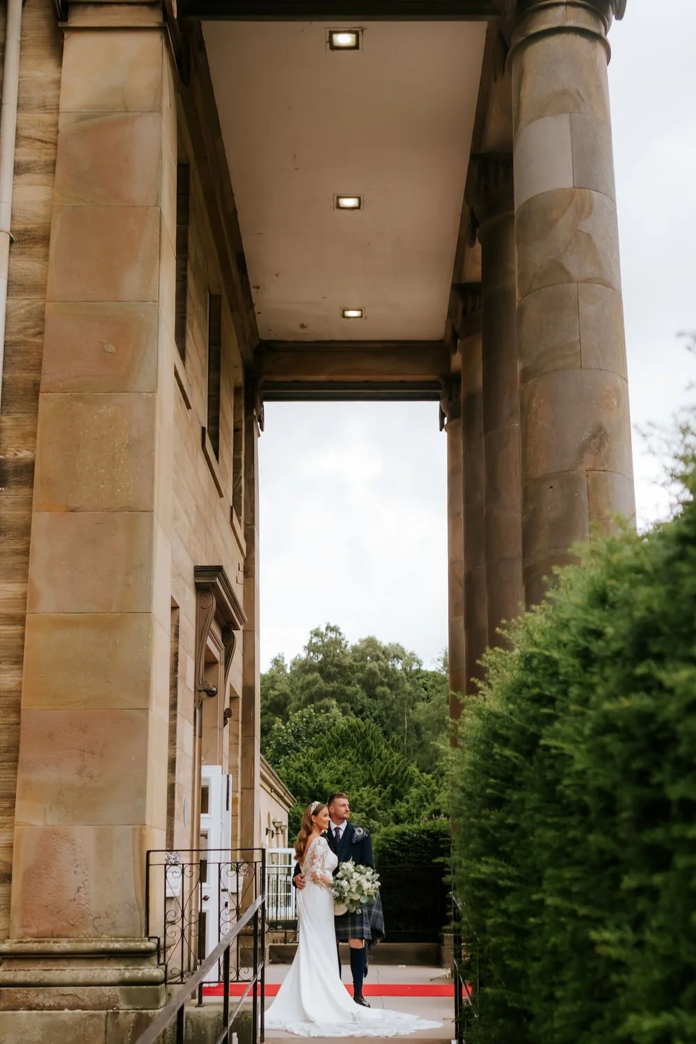 A bride and groom in wedding attire standing under a large stone archway with trees and greenery in the background.