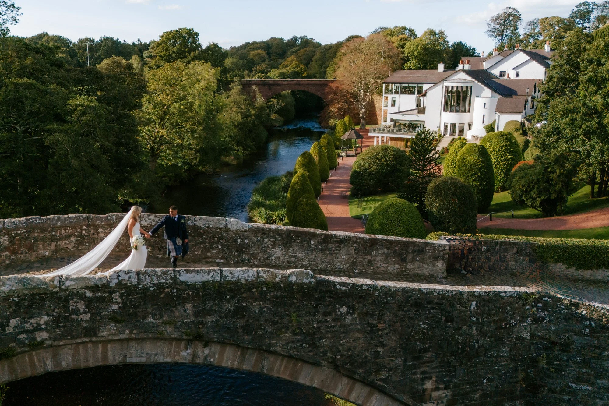 A bride and groom holding hands on a stone bridge overlooking a river, surrounded by lush greenery and a large white house in the background.