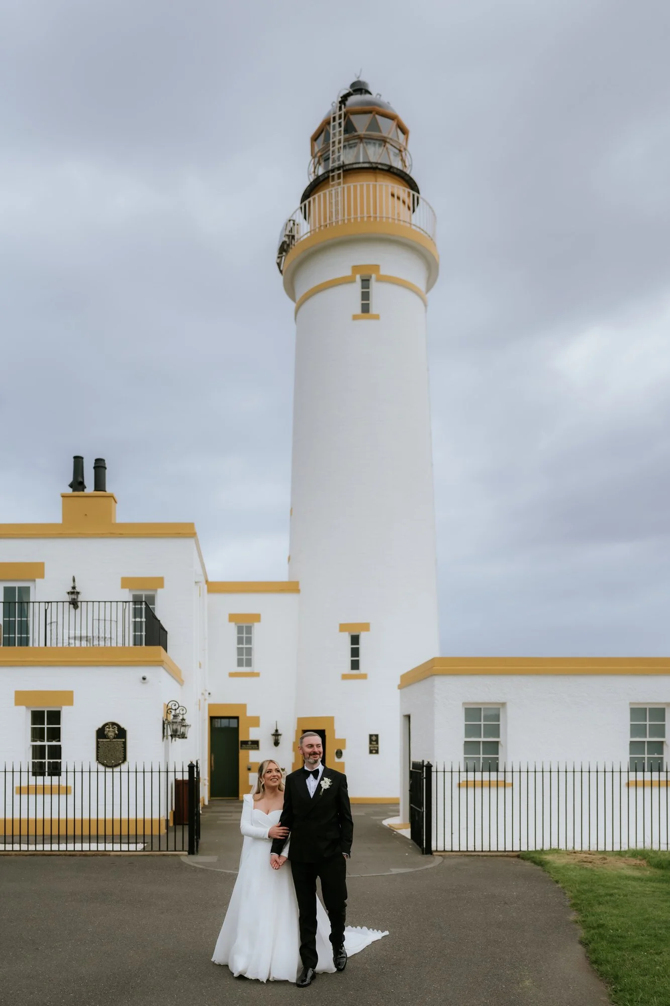 A bride and groom standing together in front of a lighthouse on a cloudy day, smiling.