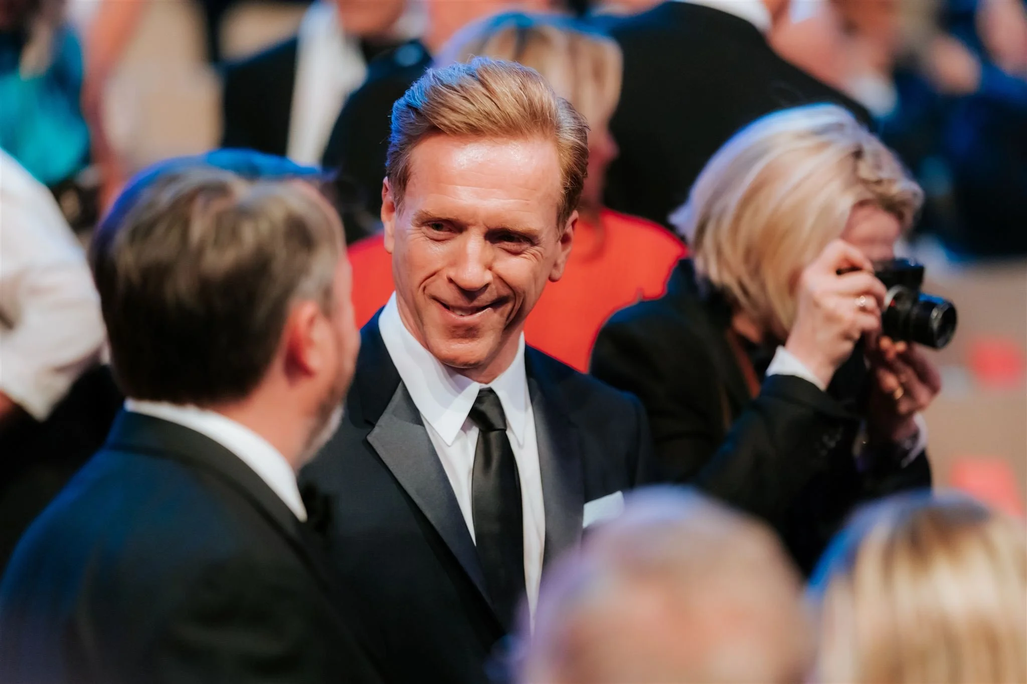 Man in a tuxedo smiling and talking to another man in a tuxedo, with a woman taking a photo nearby, at a formal event or gathering.