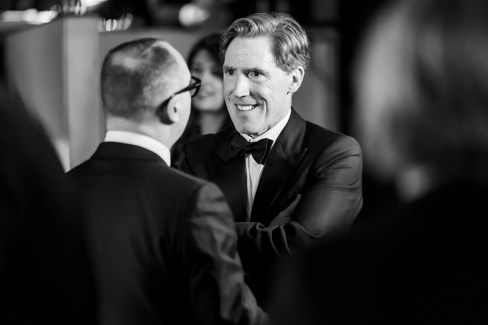 A black and white photo of two men in tuxedos engaged in conversation at a formal event, smiling at each other.