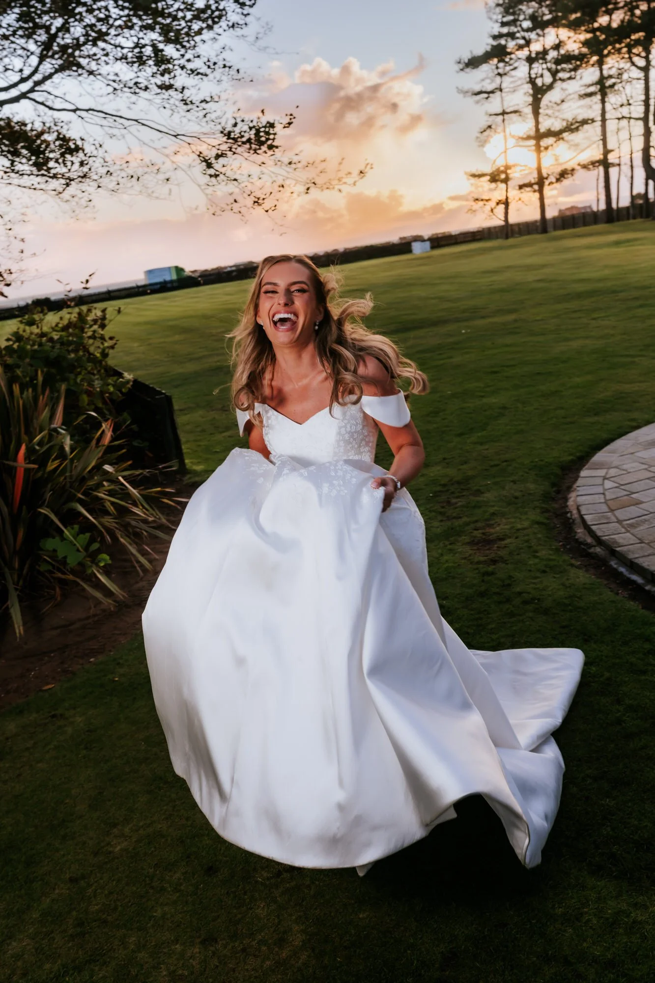 A woman in a wedding dress laughing and lifting her gown outdoors during sunset, with green grass, trees, and a cloudy sky in the background.
