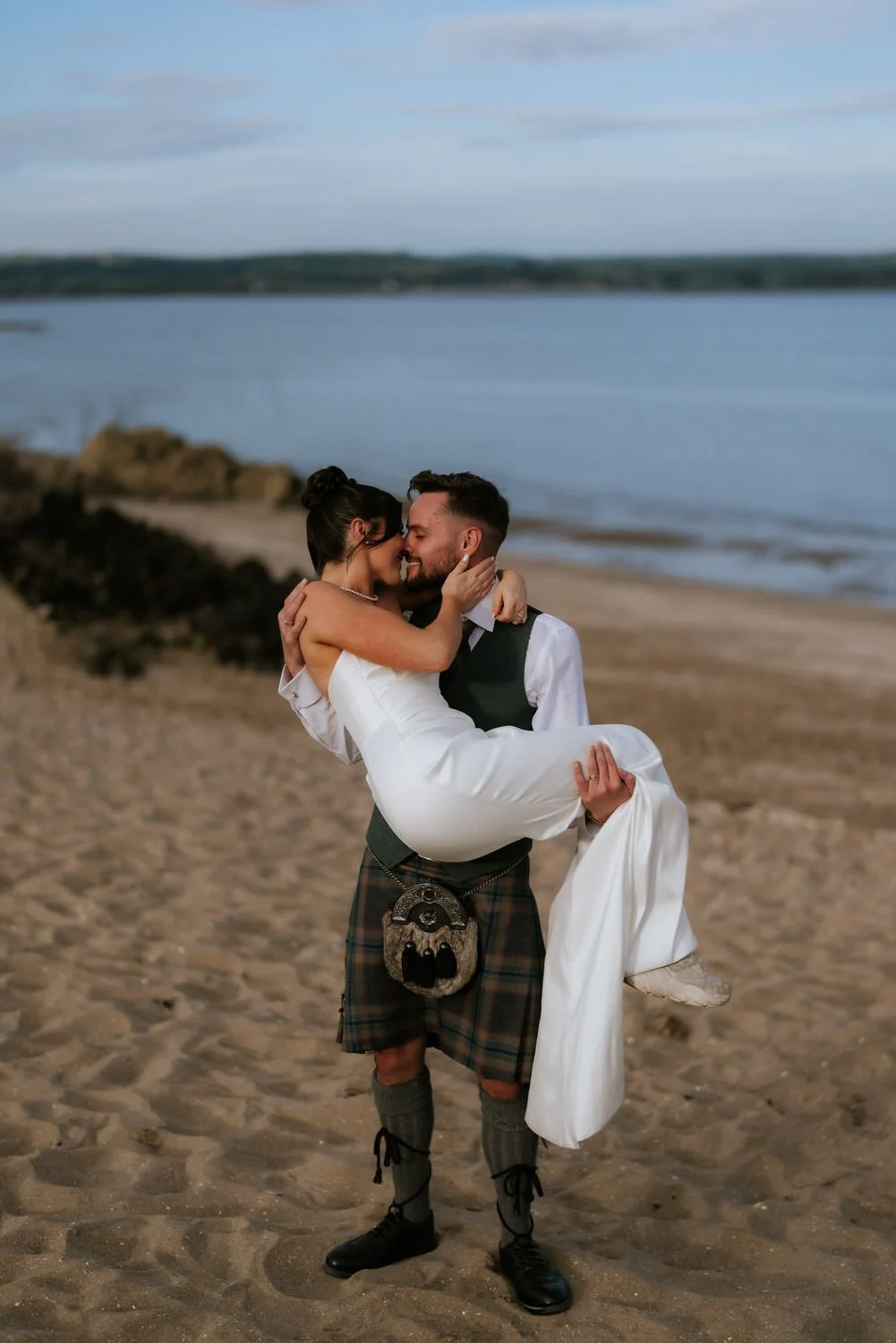 A couple dressed in wedding attire embracing on a sandy beach by the water, with one person being carried by the other.