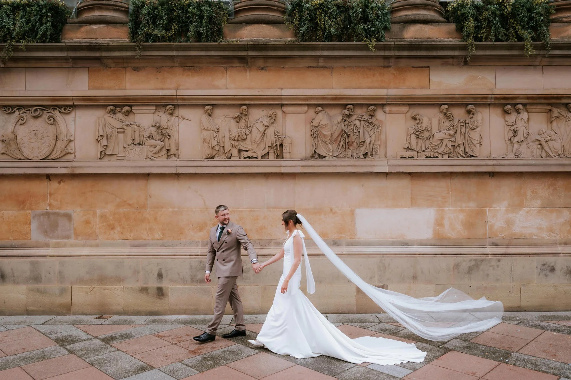 A bride and groom holding hands and walking outside against a stone wall with classical carved reliefs, the bride wearing a white gown with a long train and veil, and the groom in a brown suit.