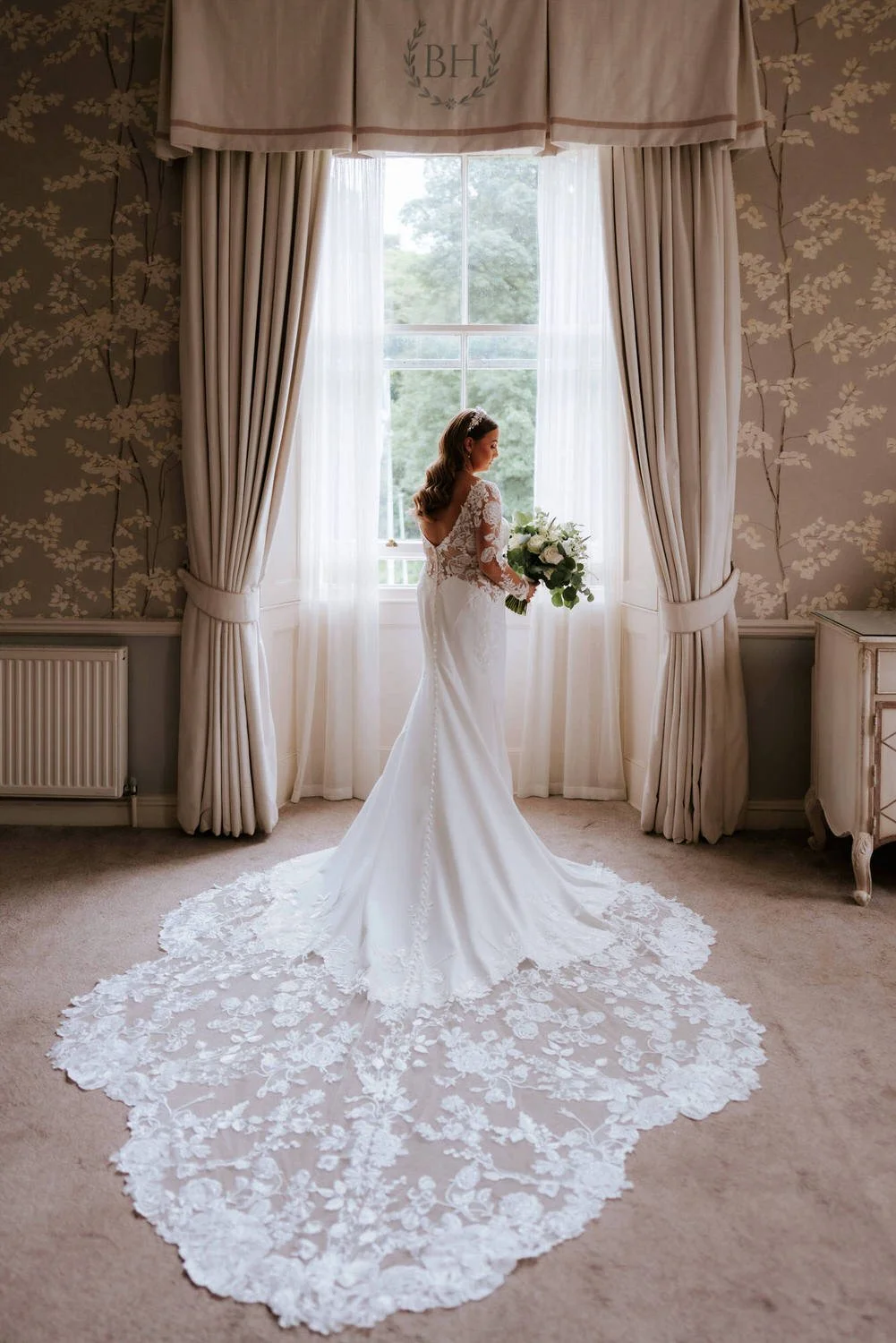 Bride in a wedding dress holding a bouquet of flowers standing by a large window in a decorated room.