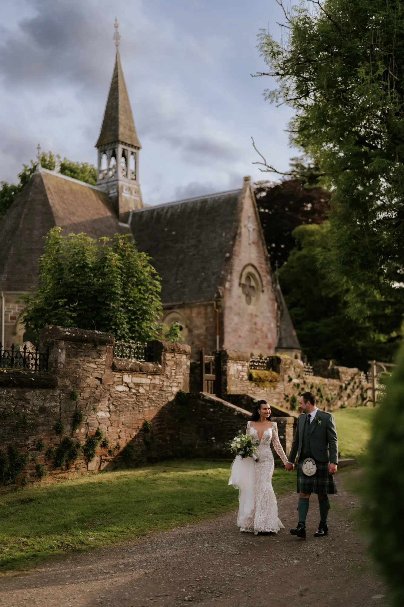 A bride and groom holding hands and walking along a dirt path outside a church, with lush greenery and trees surrounding them.