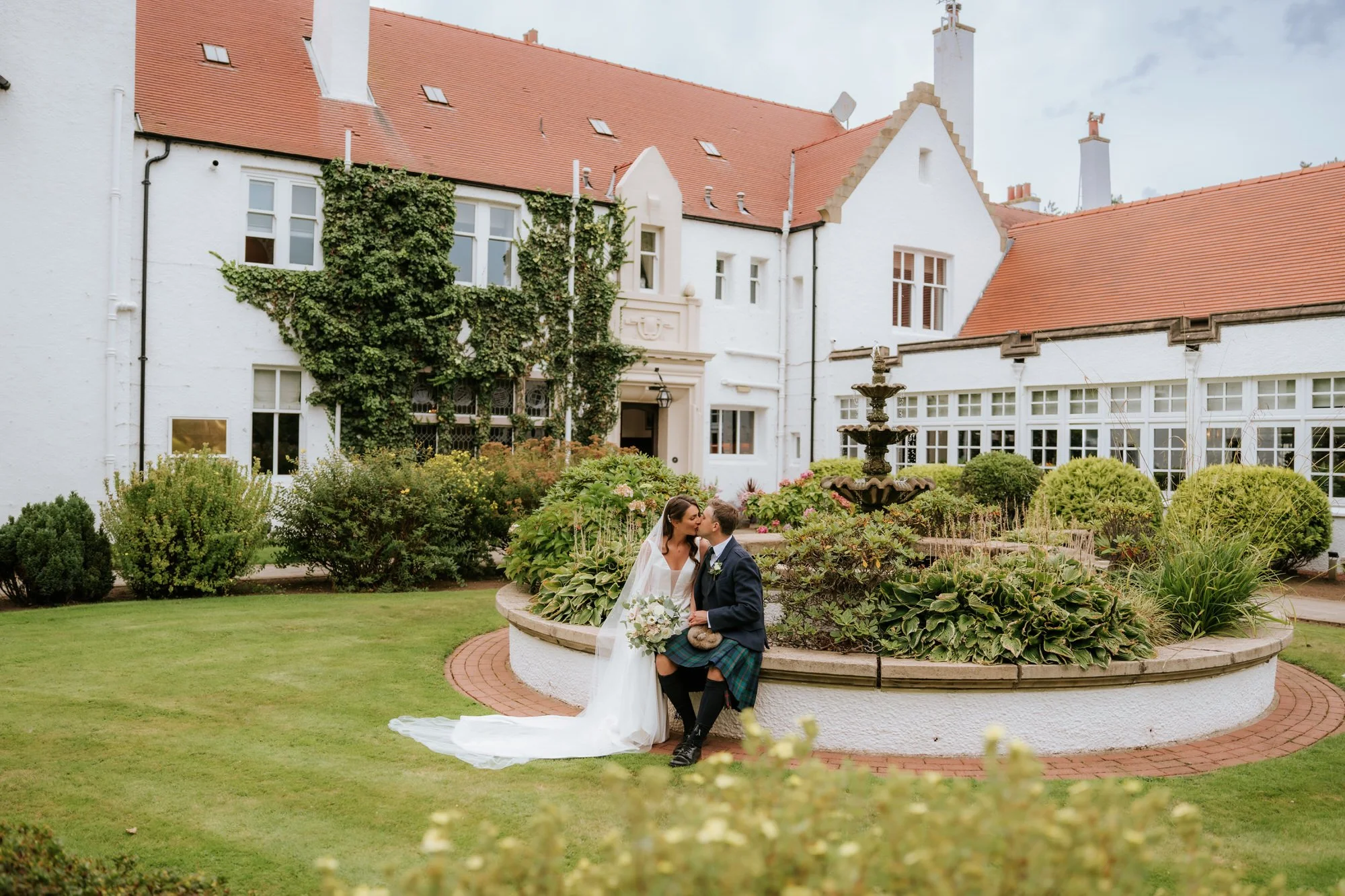 A bride and groom sharing a kiss in a garden courtyard with a fountain, green bushes, and a white building in the background.