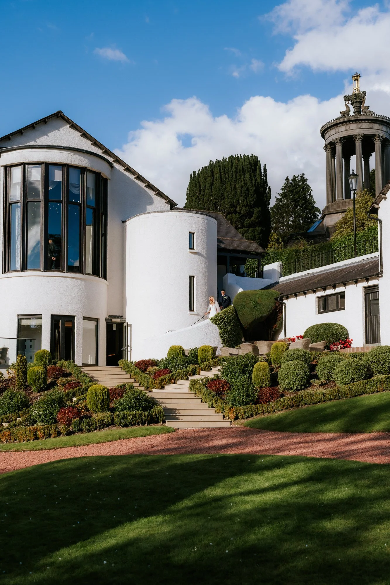 Bride and groom walking up steps outside white modern building with landscaped garden, cloudy blue sky in the background.