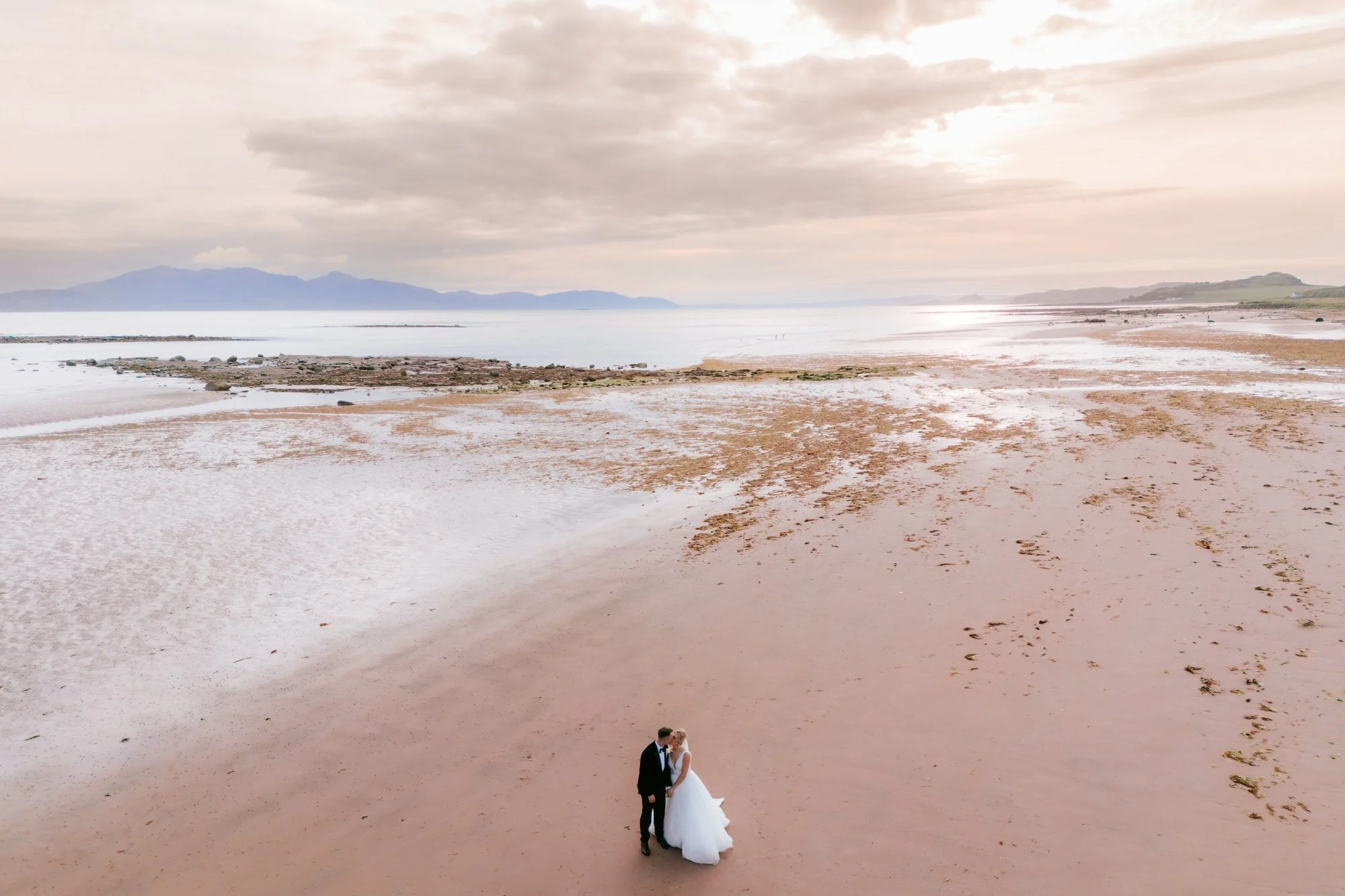 A bride and groom standing together on a sandy beach, with the ocean and a cloudy sky in the background.
