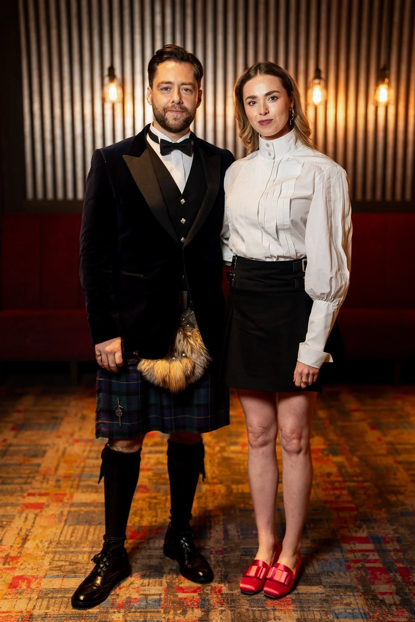 A man dressed in a tuxedo and kilt standing next to a woman wearing a white blouse and black skirt, in an indoor setting with warm lighting.
