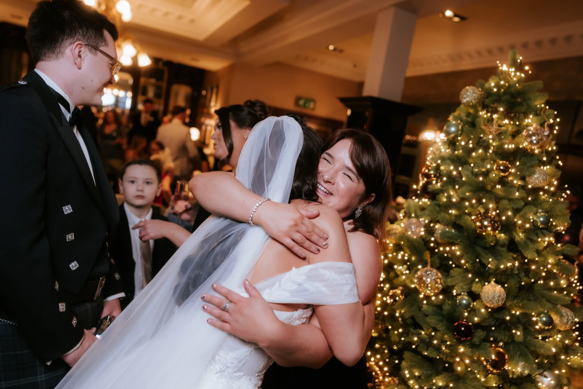 Bride hugging a smiling wedding guest beside a lit Christmas tree at Boclair House