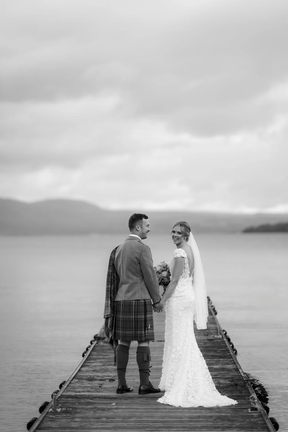 A black and white photo of a wedding couple standing on a dock over water, holding hands, with mountains and cloudy sky in the background.