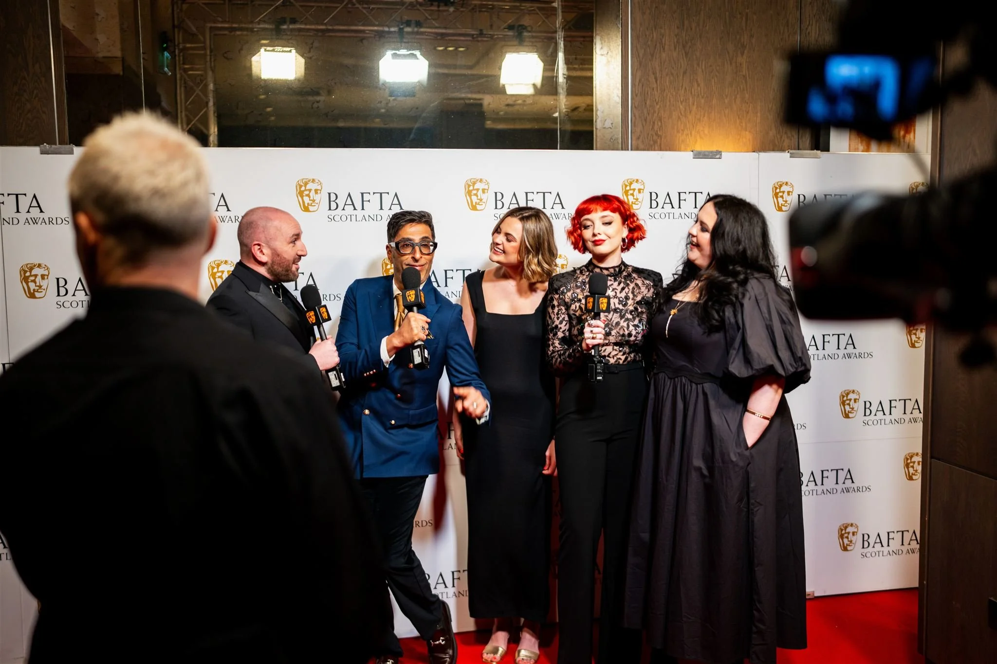 Five people, three women and two men, are standing on a red carpet at the BAFTA Scotland Awards, being interviewed by a host holding a microphone. A large BAFTA Scotland Awards backdrop is behind them. The scene is illuminated with bright lights, and